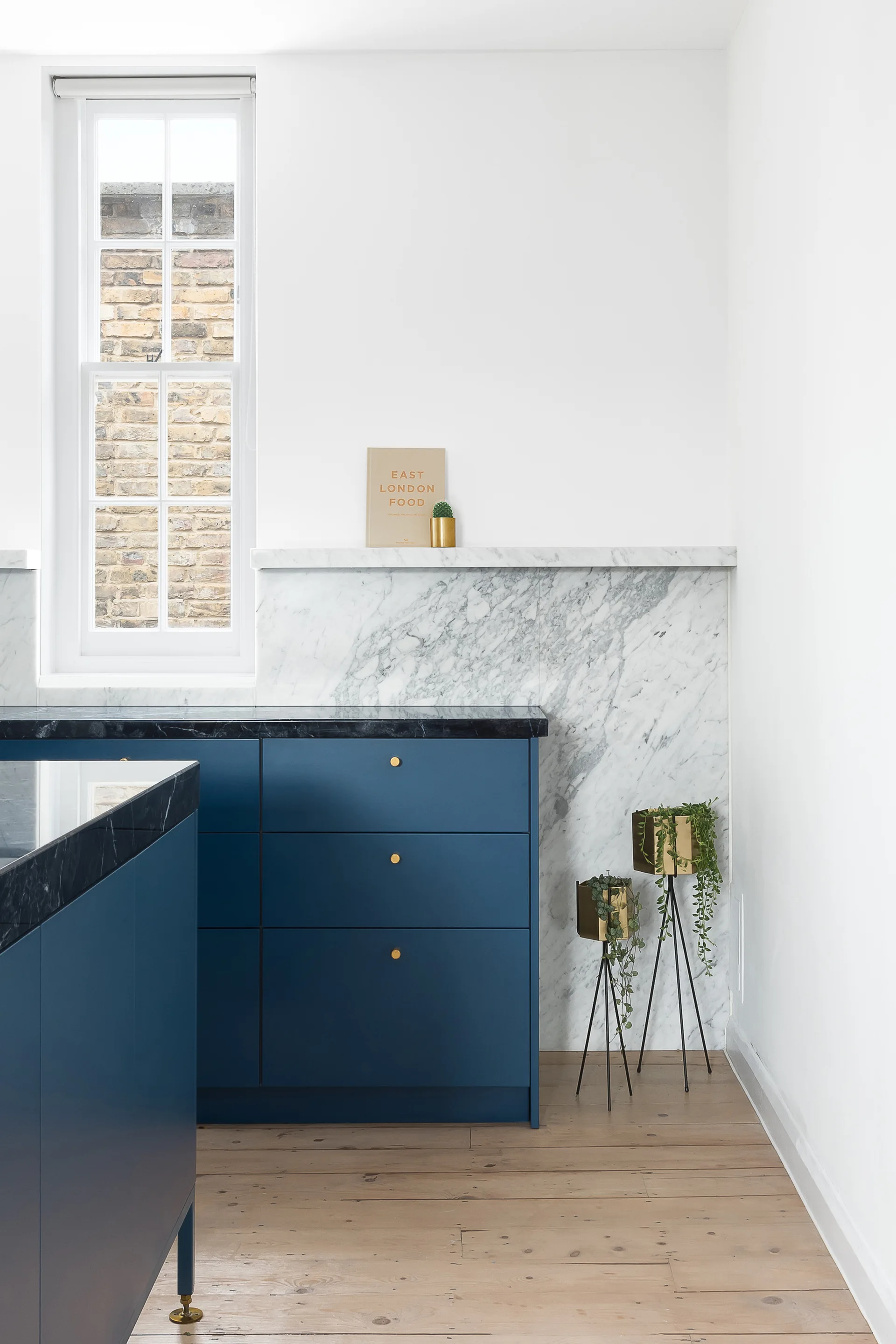 Deep blue kitchen cabinets feature small brass knobs, paired with a white marble slab backsplash, dark countertop, and light wood floors.