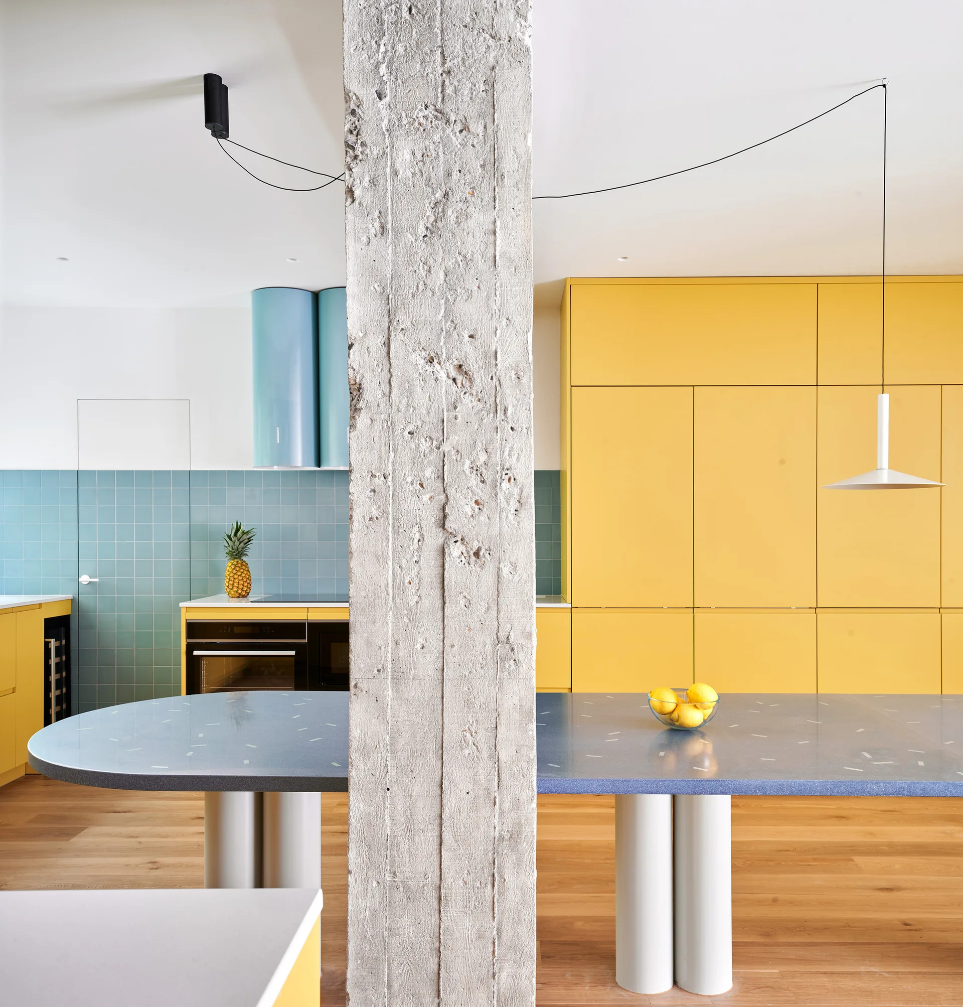 A modern kitchen with tall yellow cabinets, a blue tiled backsplash, and a blue island positioned behind a rough, exposed concrete column.
