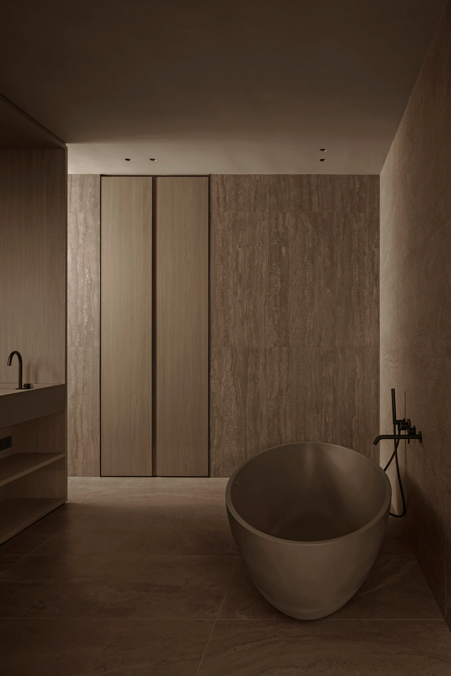 A minimalist bathroom featuring an oval freestanding tub, matte black wall-mounted faucet, and warm brown stone wall tiles next to integrated wood cabinets.