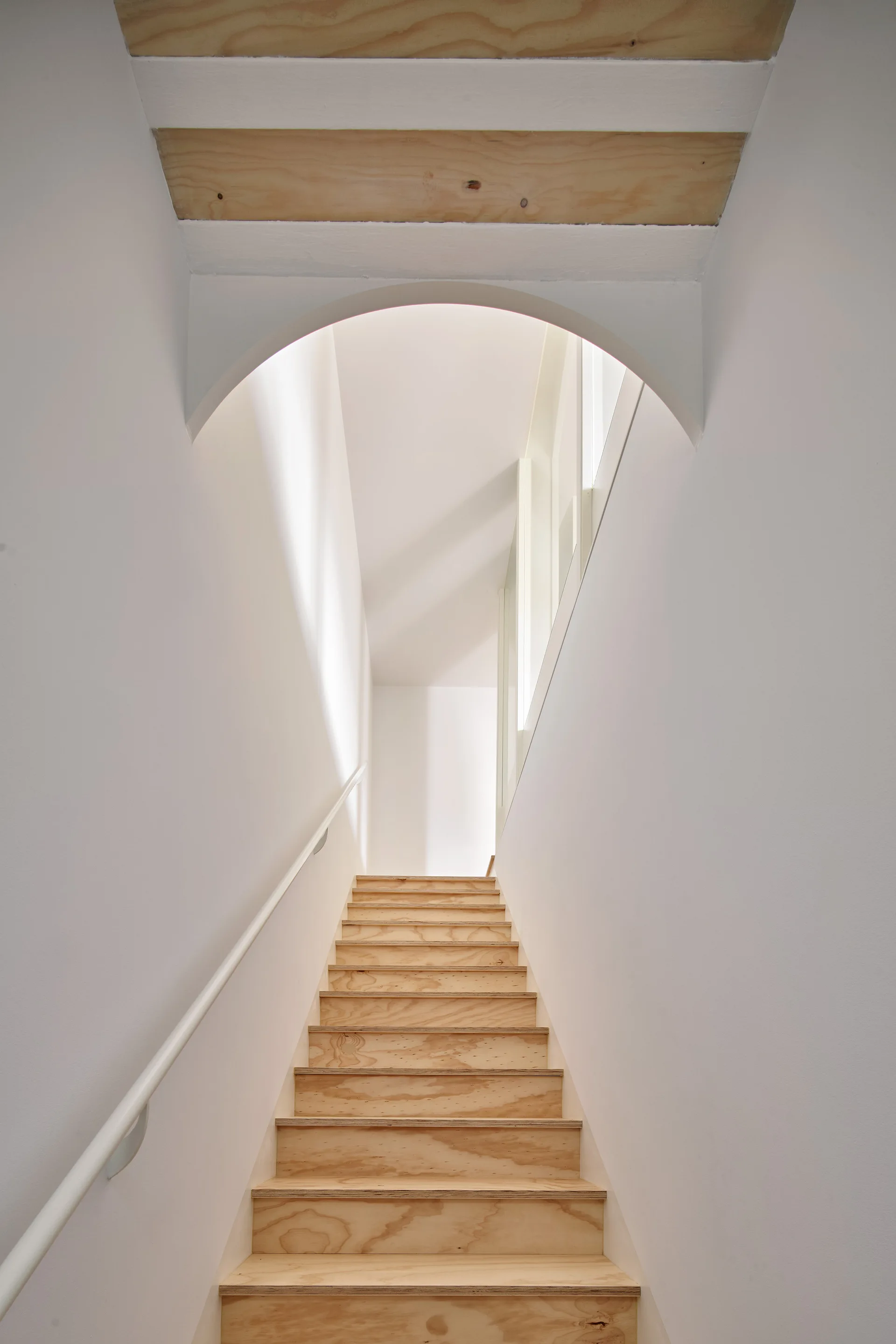A minimalist wooden staircase with light-toned treads ascends between white walls under a smooth white plaster archway.