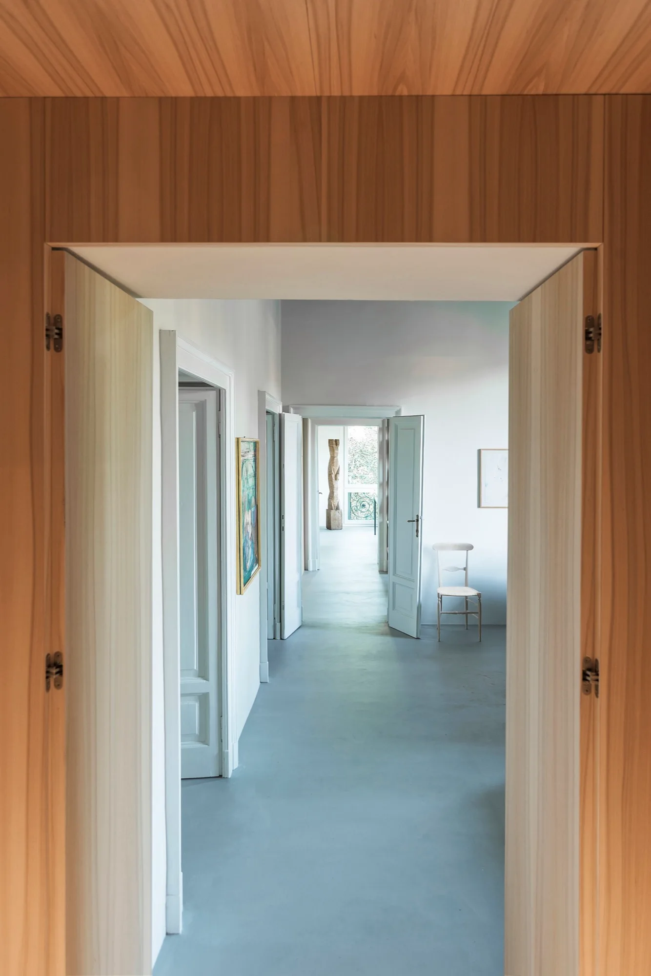A view from a wood-paneled doorway looks down a long white hallway with open doors, grey floors, a single chair, and a distant sculpture.