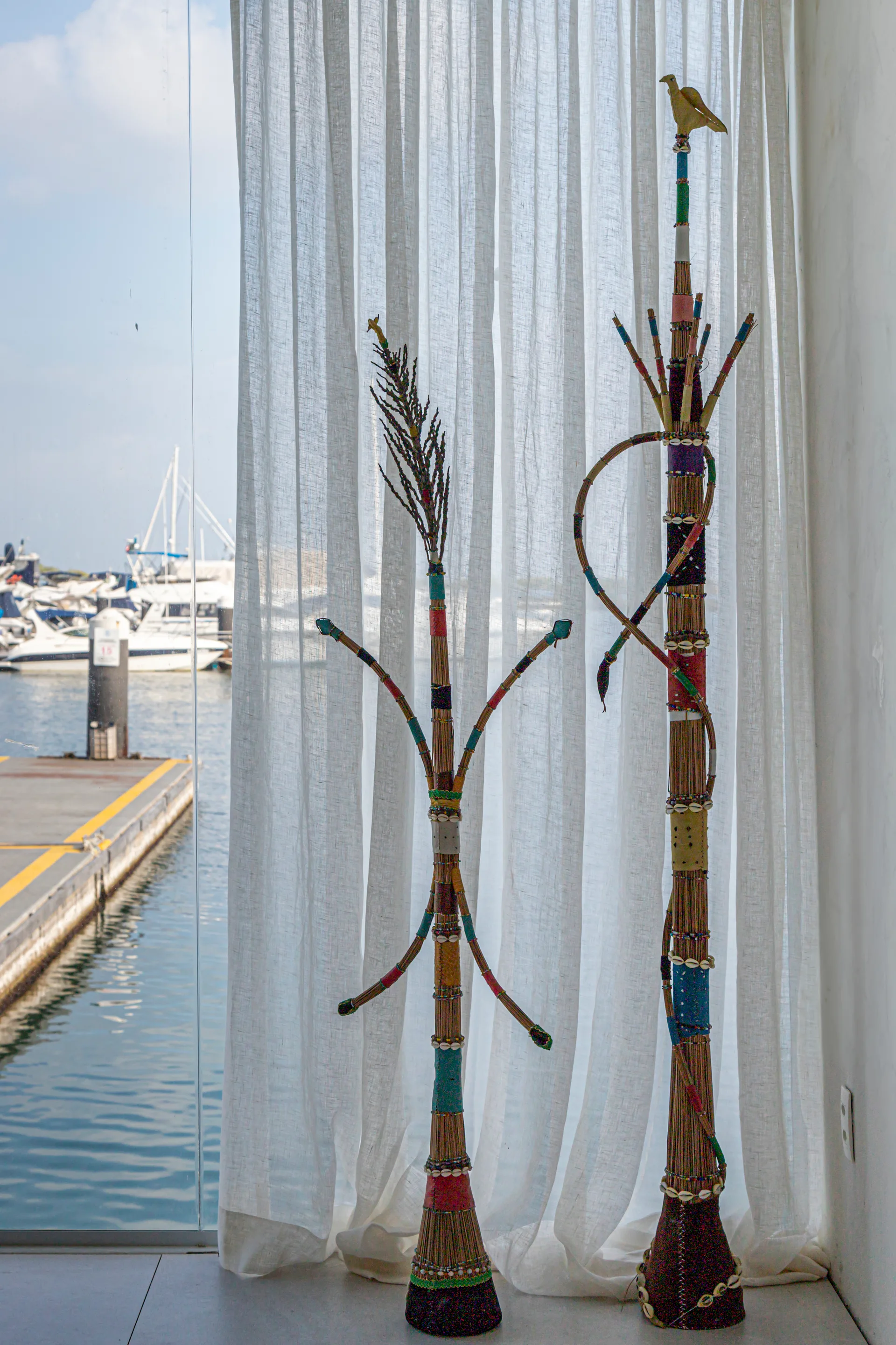 Two tall, handcrafted fiber sculptures adorned with beads and shells stand before a sheer white curtain overlooking a marina.