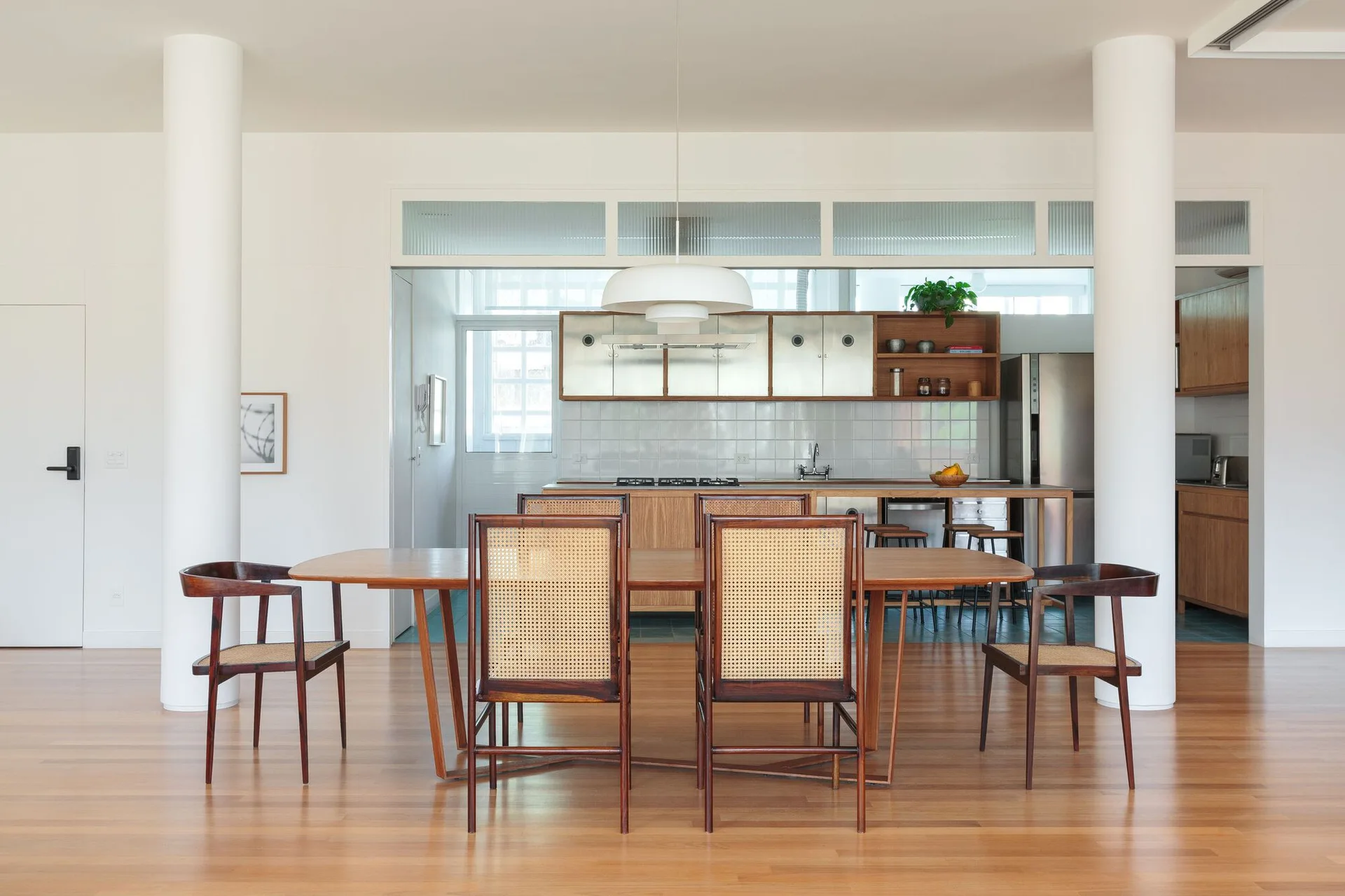 A wooden dining table with woven cane chairs stands between two white columns, with an open kitchen featuring metallic cabinets visible in the background.