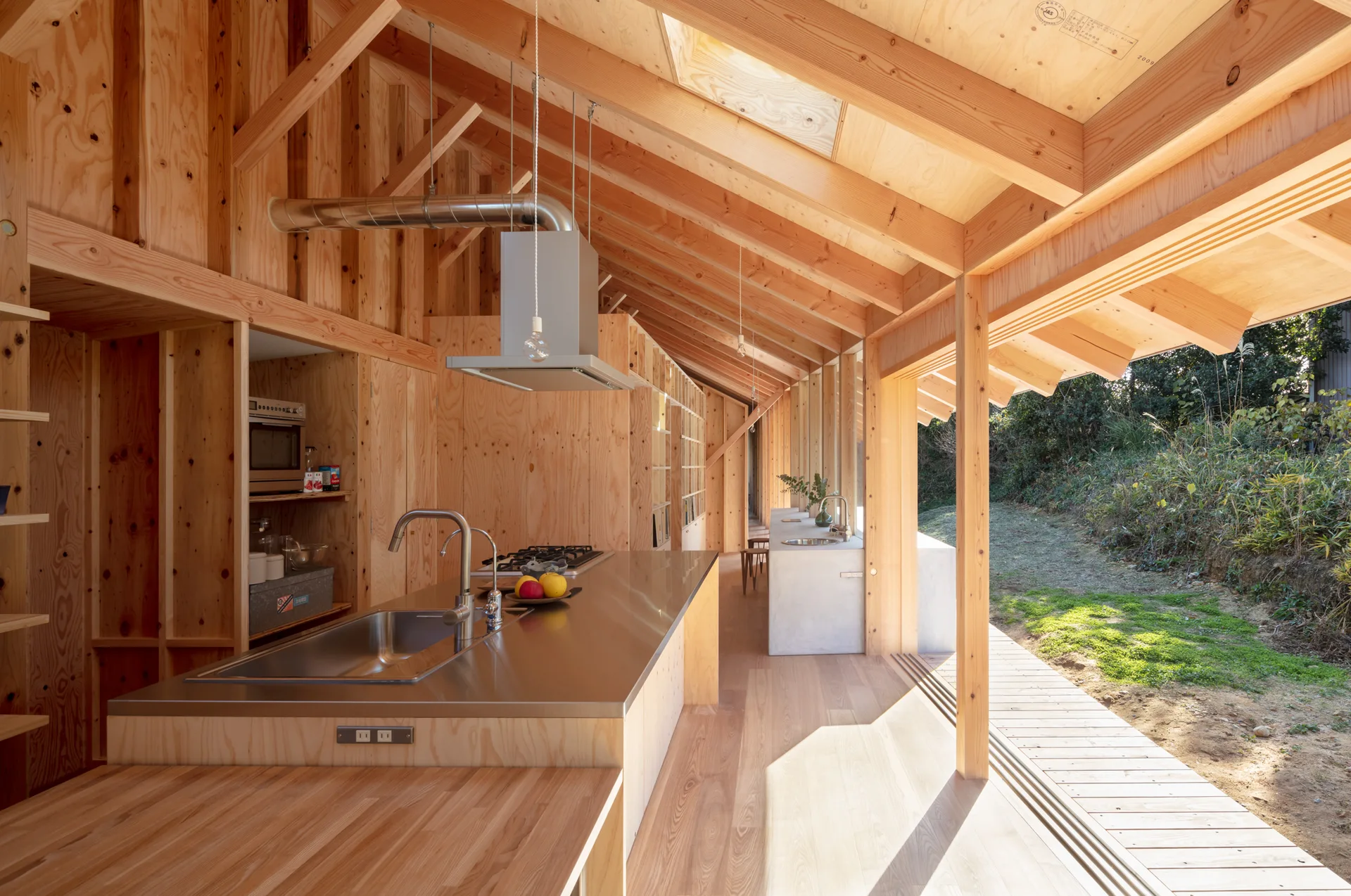 A minimalist wood-paneled kitchen features a long stainless steel island, exposed rafters, and sliding glass doors opening to an outdoor deck.
