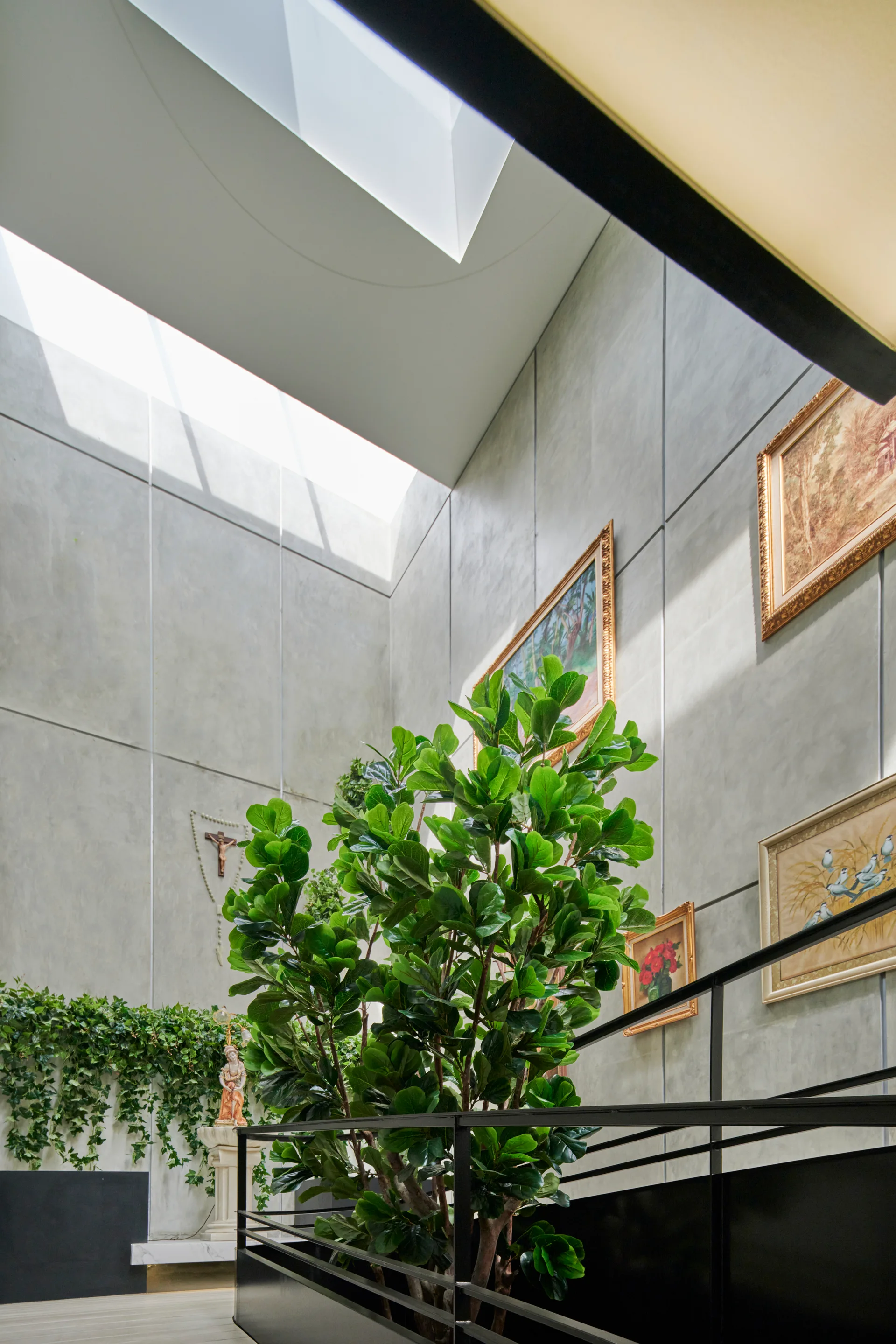 A tall staircase atrium features concrete panel walls with gold-framed paintings, a black metal railing, and a large leafy green plant under a skylight.