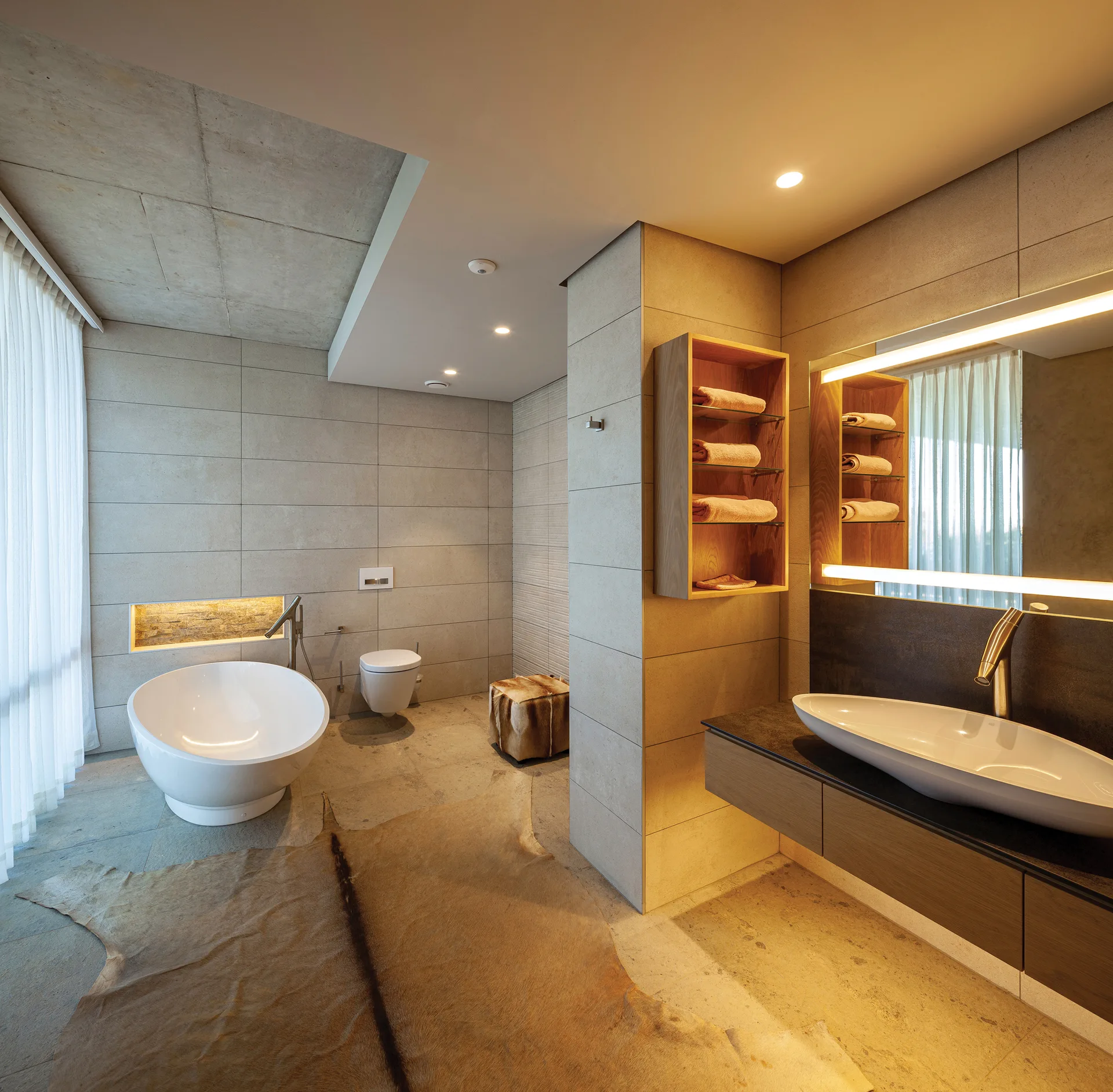 A modern bathroom clad in beige stone tiles features a round freestanding tub, a floating vanity with a vessel sink, and a large hide rug on the floor.