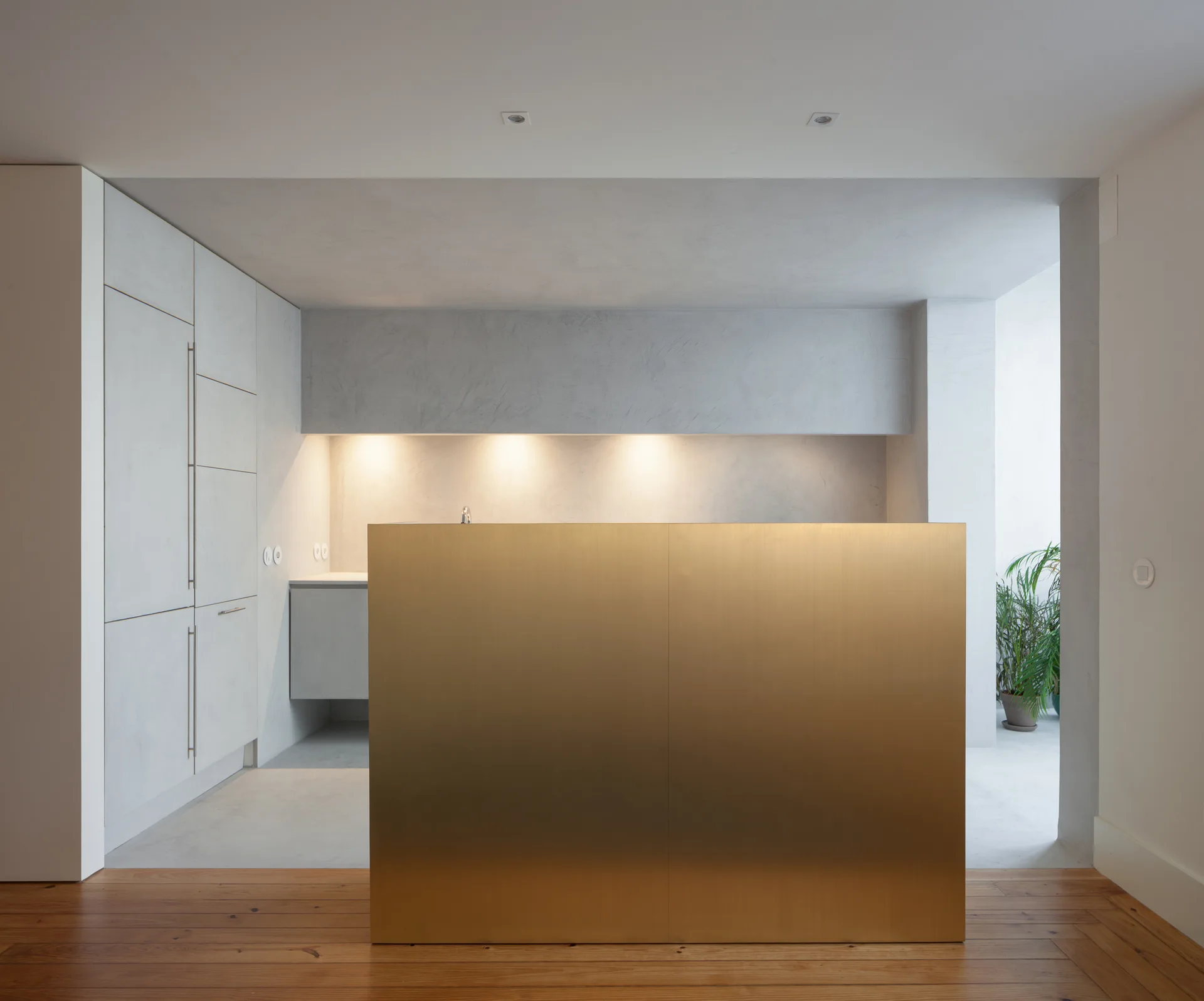 A minimalist kitchen with a large brass-faced island, light gray concrete-look cabinetry, and warm wood flooring in the foreground.