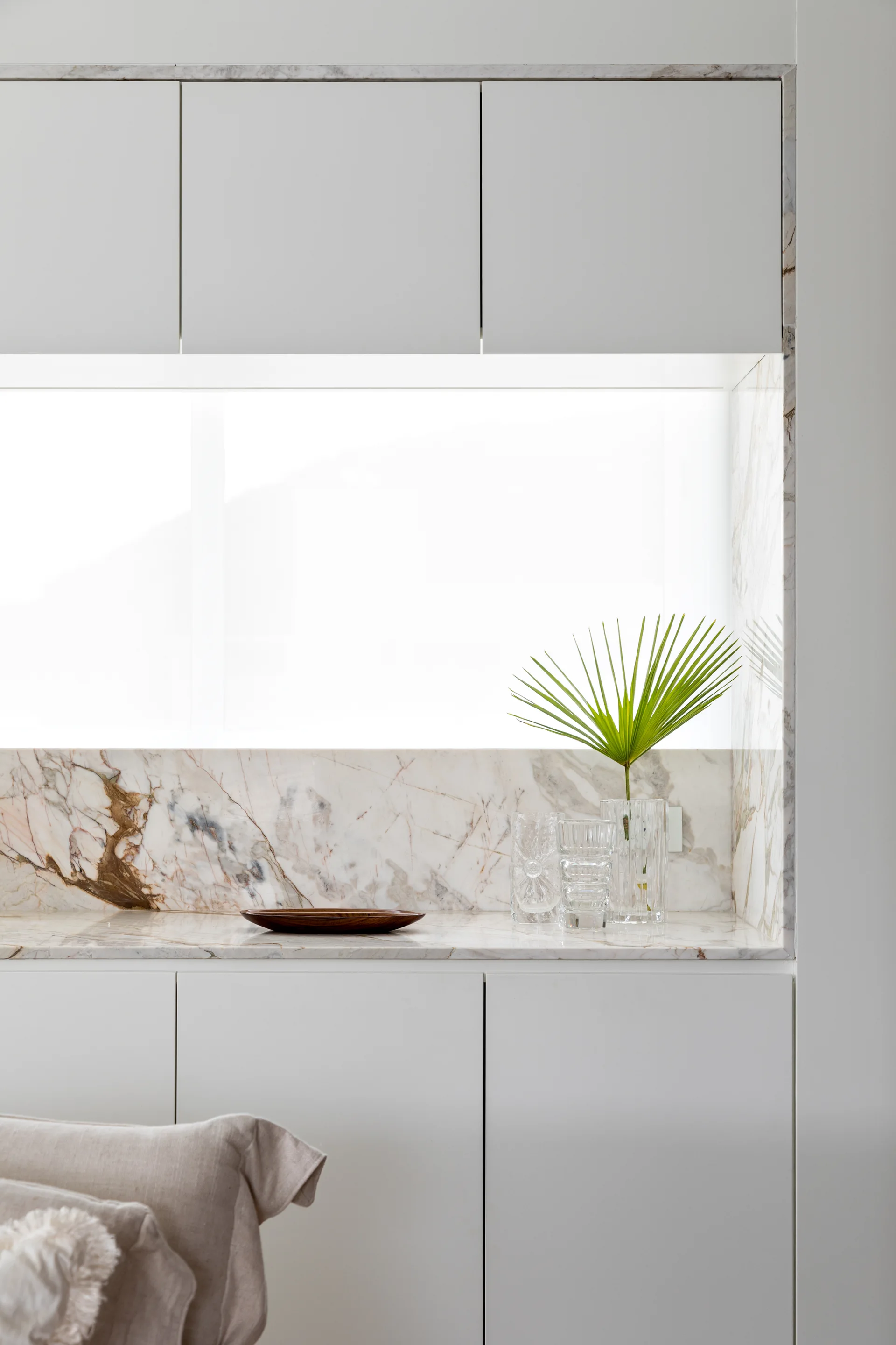 A close-up of a minimalist kitchen counter with matte white flat-panel cabinets, a veined marble backsplash, a small wooden dish, and clear glass vases.