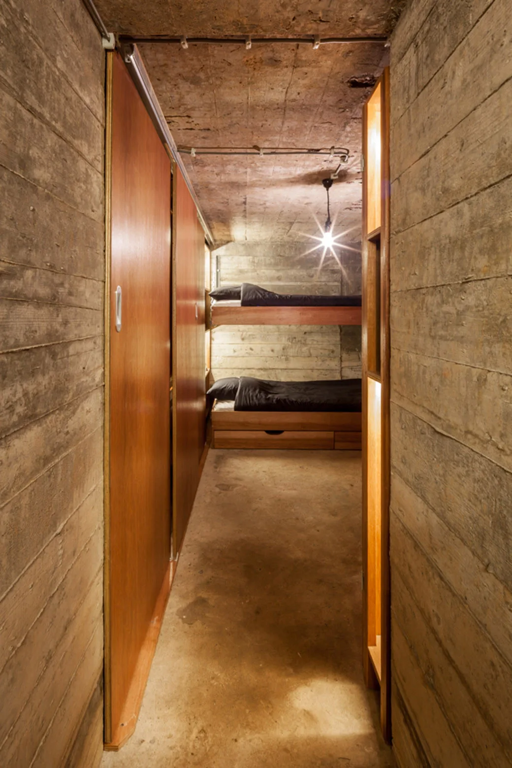 A narrow concrete bedroom featuring wooden sliding doors, board-formed concrete walls, and bunk beds with black bedding.