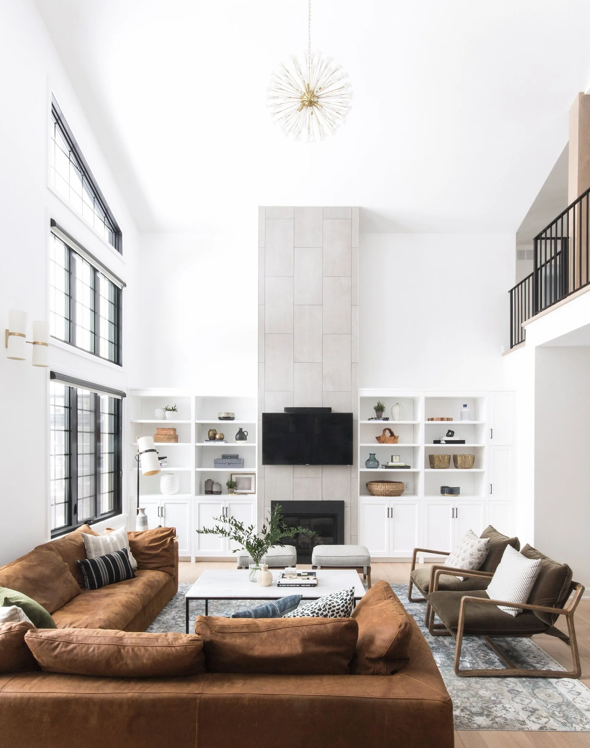 A bright living room with a tall tiled fireplace, symmetrical white built-in shelves, a tan leather sectional sofa, and a gold sputnik chandelier.