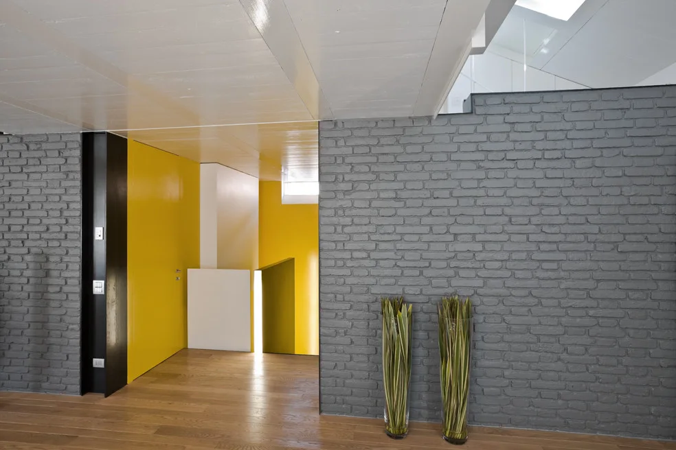 A modern hallway features a textured grey painted brick wall, a bright yellow door, and natural wood flooring under a white ceiling.