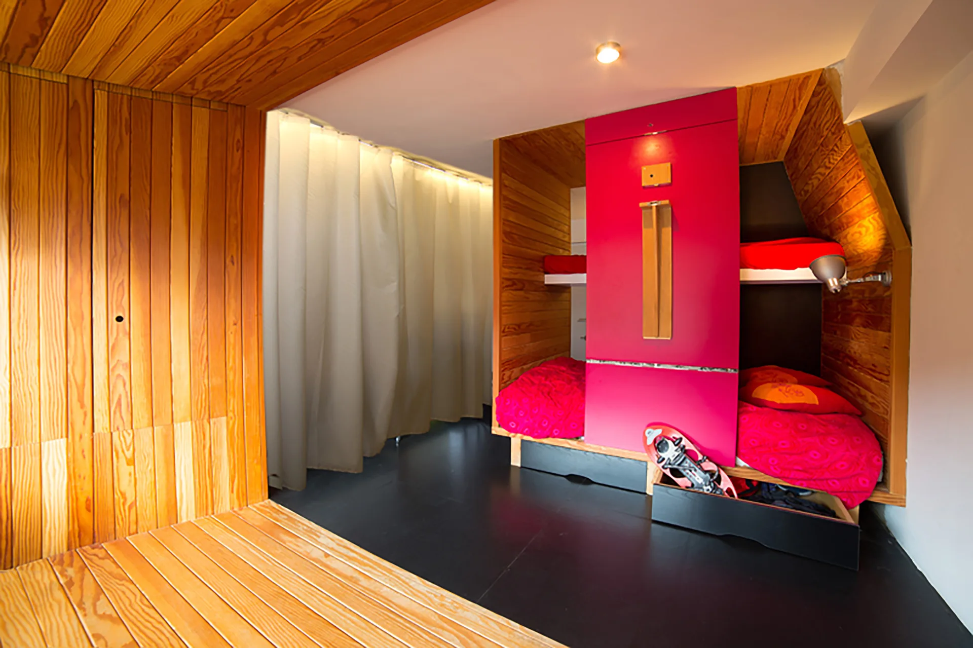 Built-in wooden bunk beds feature natural wood paneling, a vibrant hot pink storage unit, and dark flooring next to a white curtain partition.
