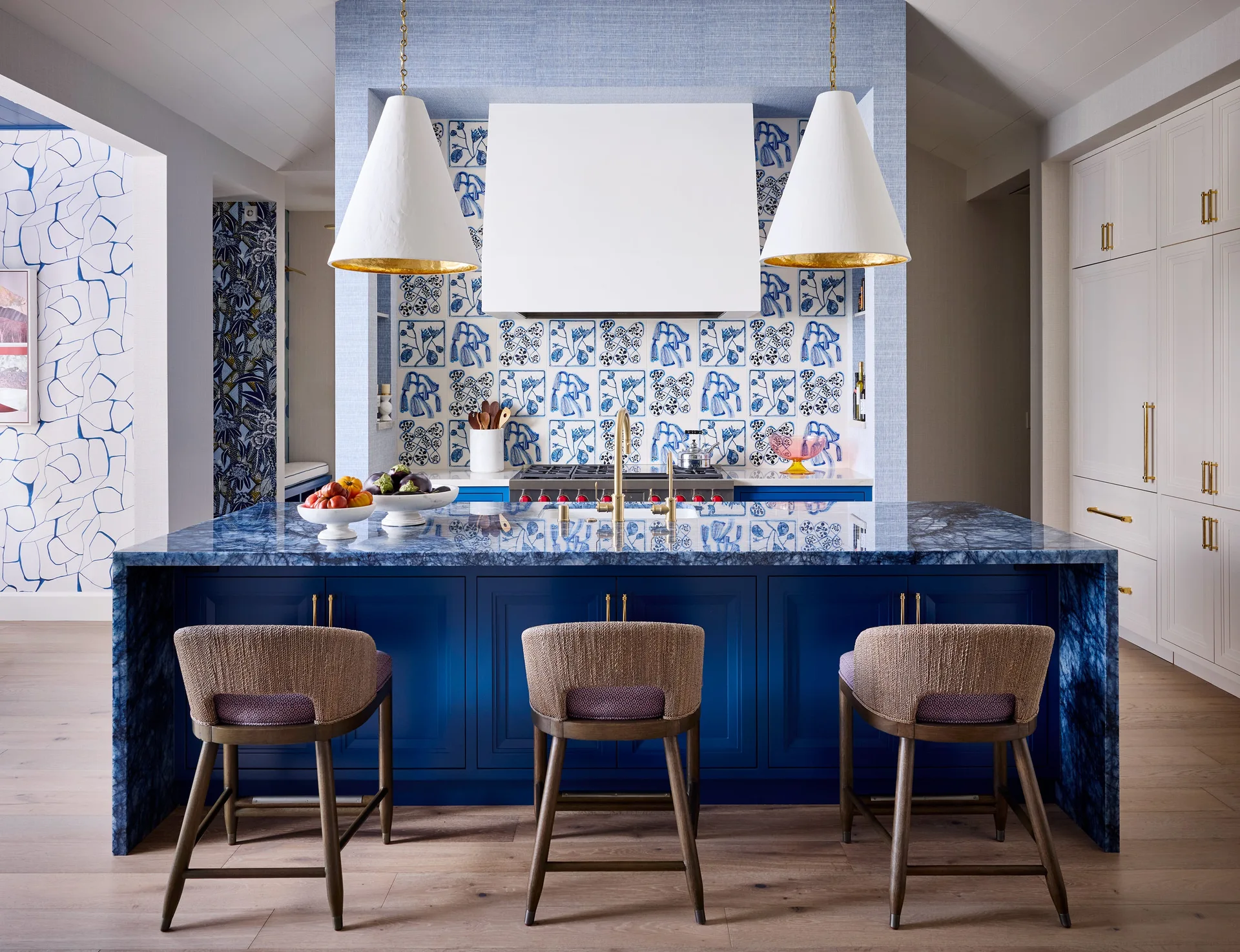 A blue marble kitchen island with three woven bar stools stands before a stove area with blue and white patterned tiles and cone pendants.