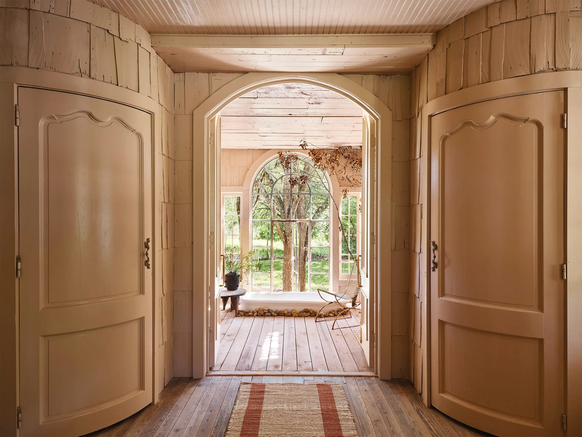 An arched doorway leads into a sunlit room with a white bathtub, rustic wood flooring, and a large arched window looking out at trees.