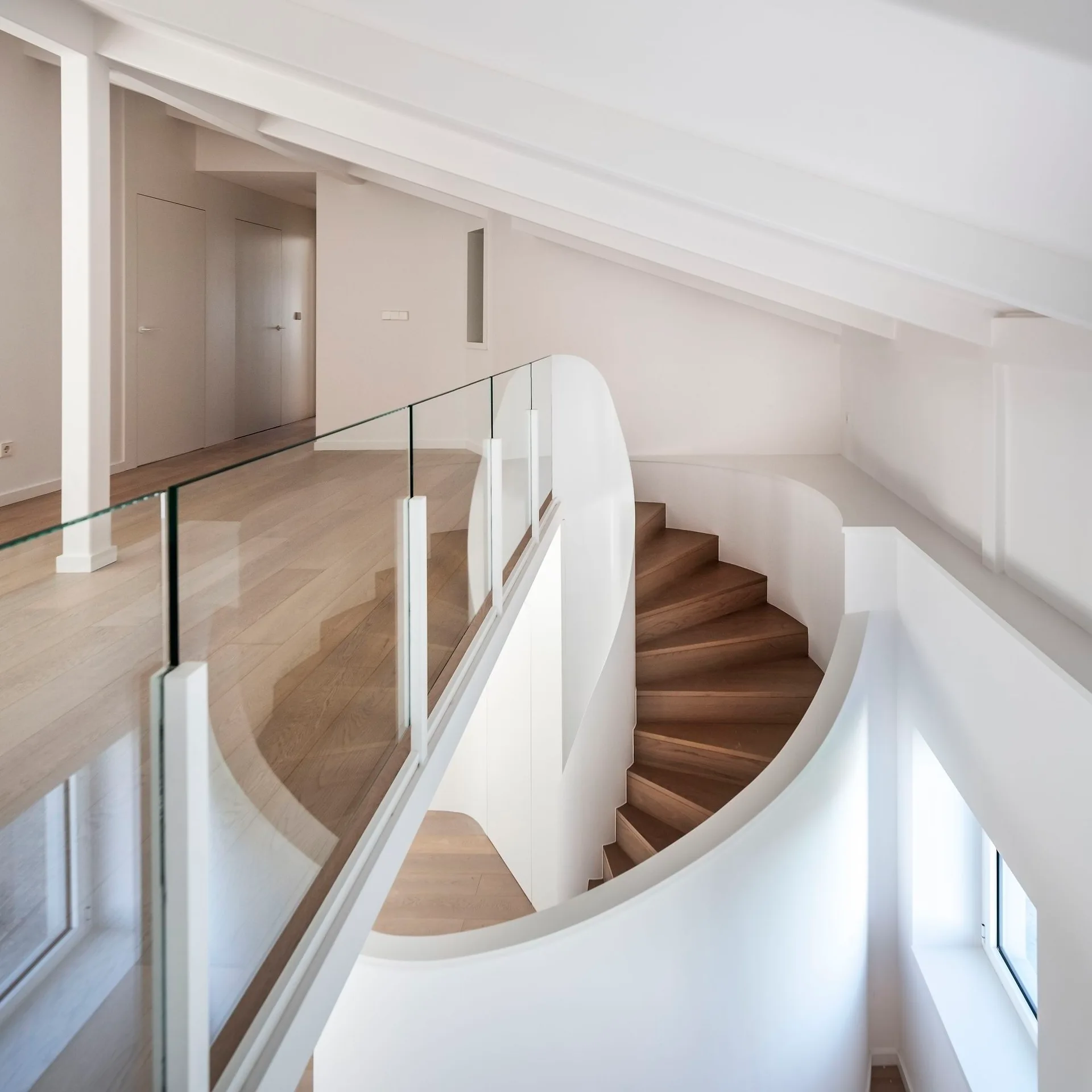 A modern loft landing features a curved wooden spiral staircase, transparent glass railings, and white sloped ceilings with exposed beams.