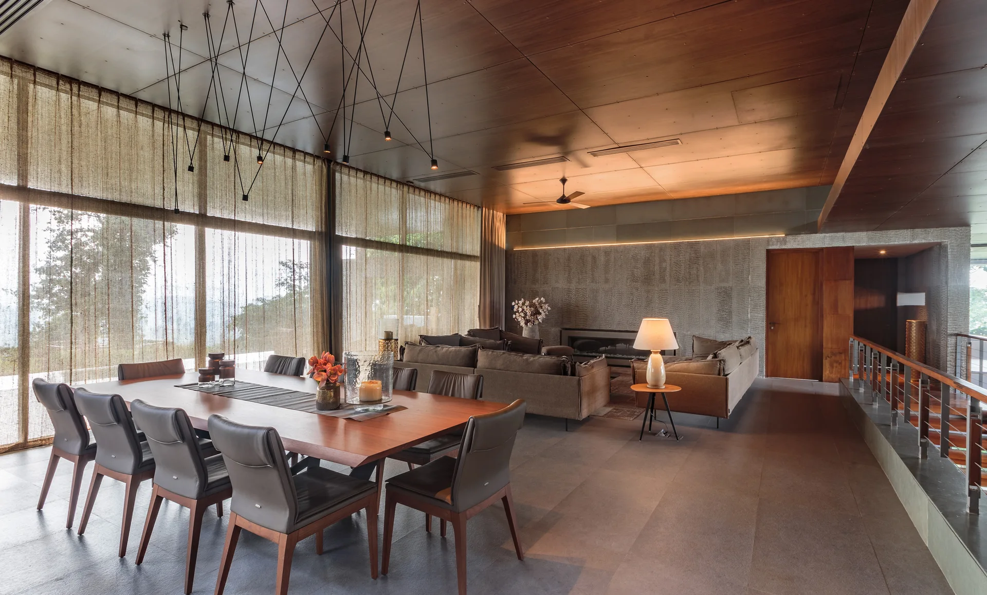 A spacious open-plan dining room features a large wooden table with leather chairs, a wood-paneled ceiling, and sheer curtains over large windows.