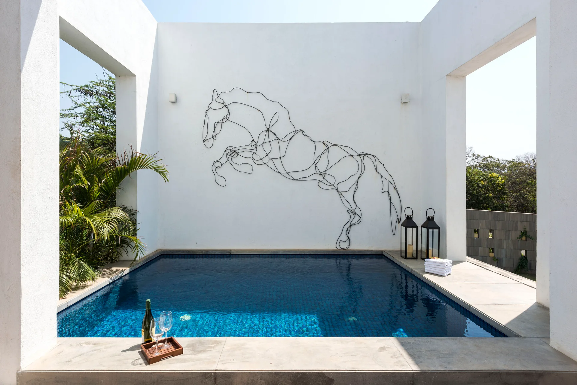 A small blue swimming pool is framed by white stucco columns and features a large black wire horse sculpture on the back wall.