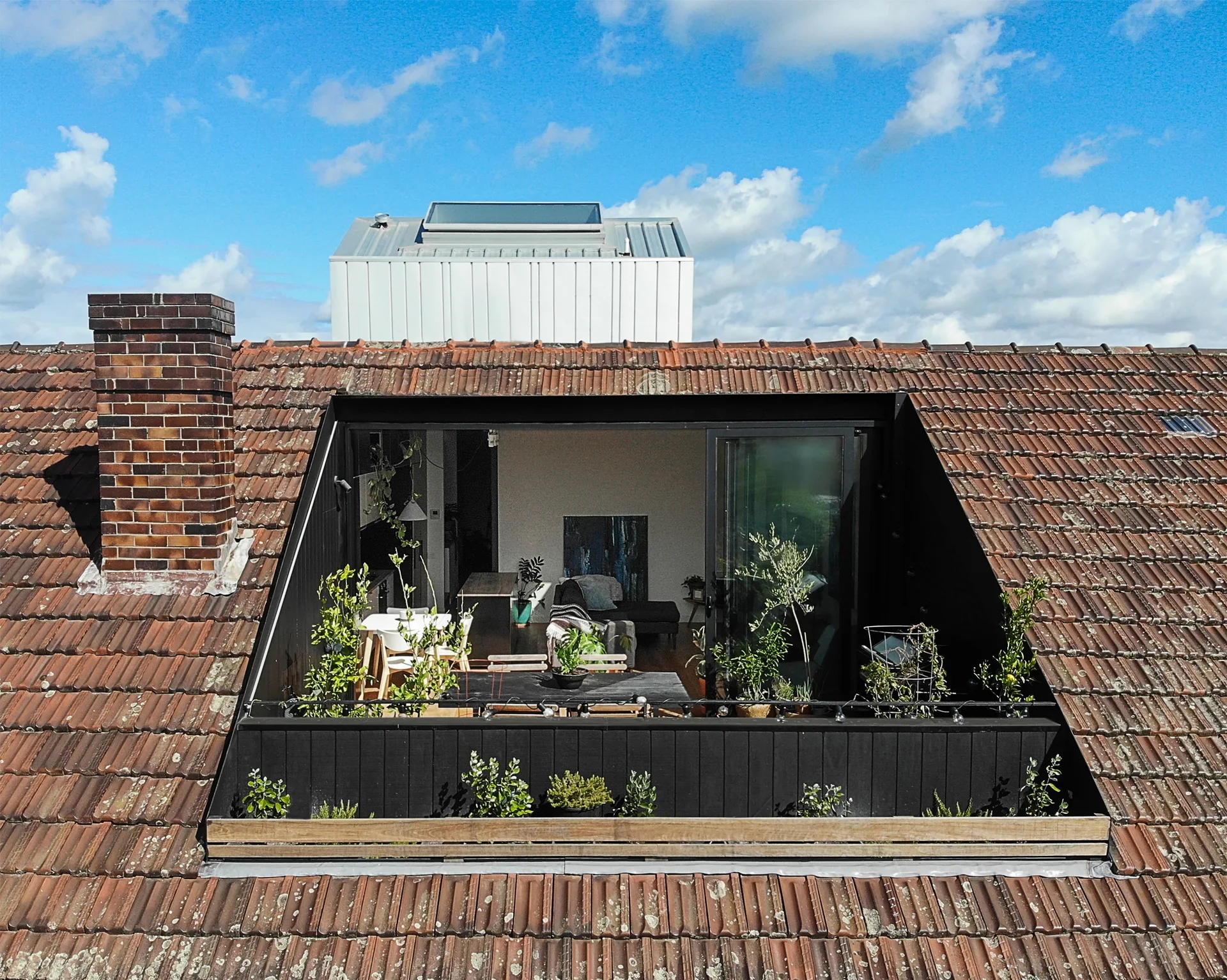 A recessed modern black balcony set within a weathered terracotta tile roof, filled with potted plants and outdoor furniture next to sliding glass doors.