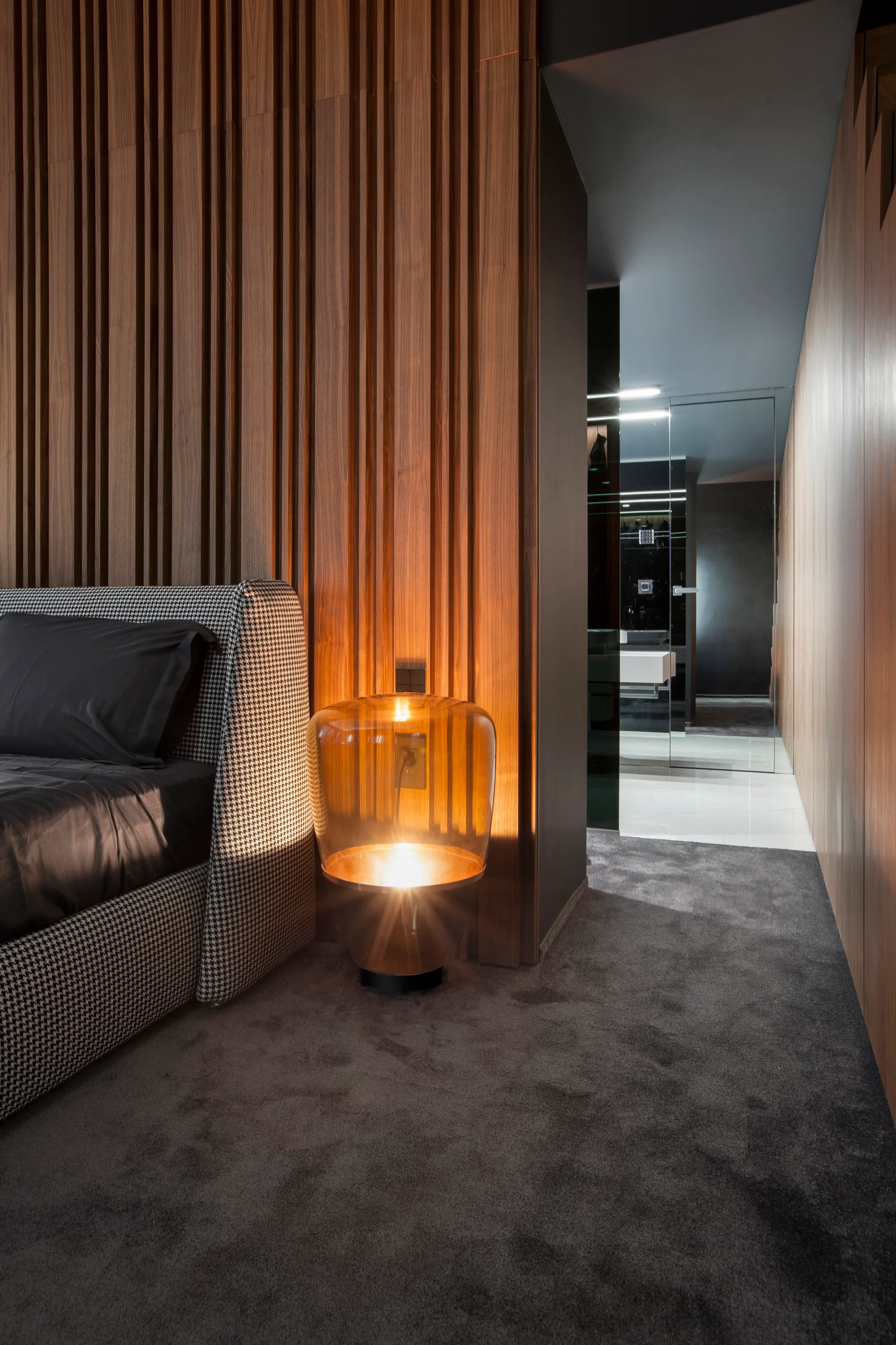 A modern bedroom features a houndstooth upholstered bed, a glowing amber glass bedside lamp, and vertical wood slat wall paneling.
