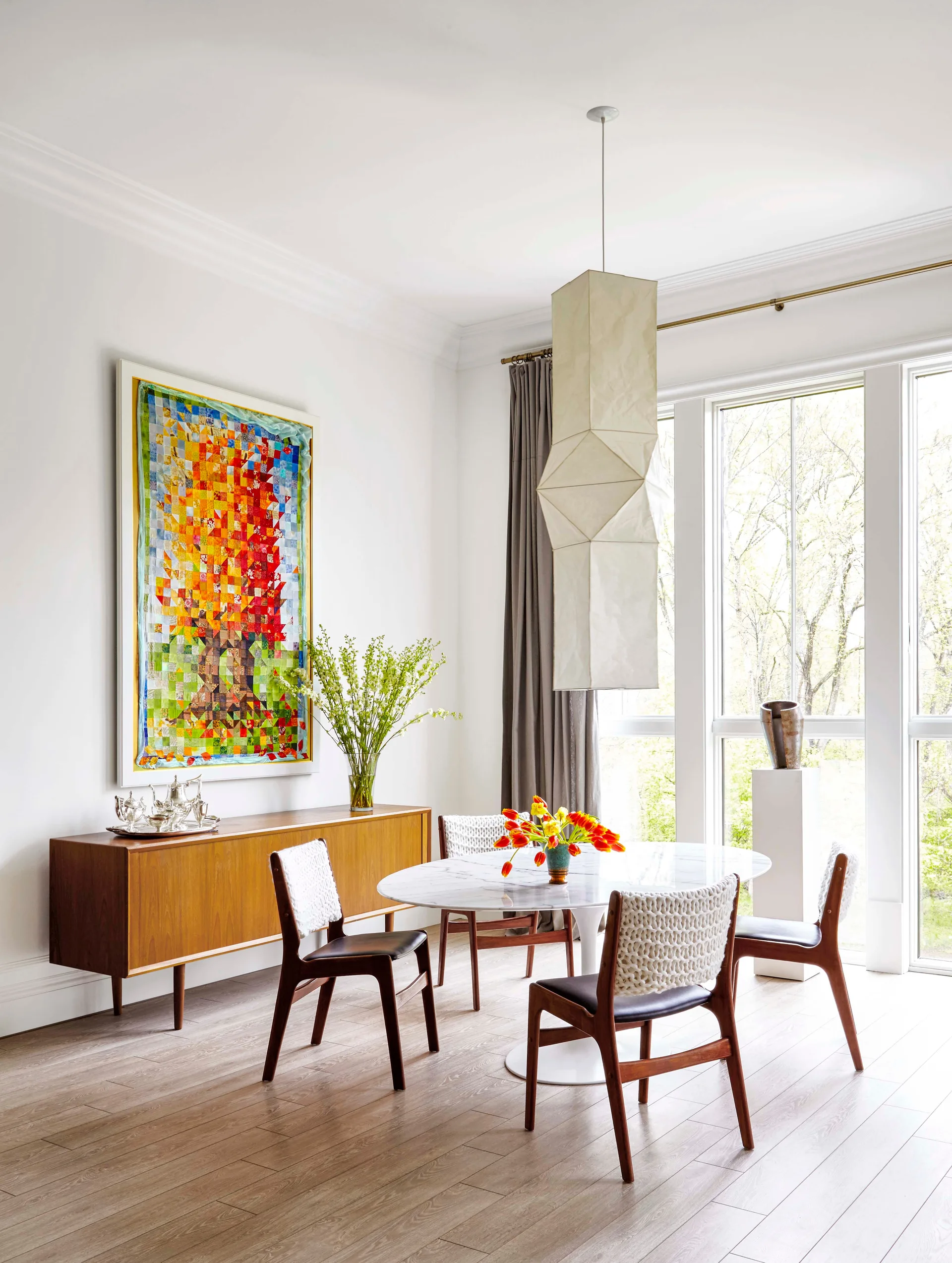 A bright dining room with an oval marble table, chairs featuring woven white backs, a wooden sideboard, and a tall paper pendant light.