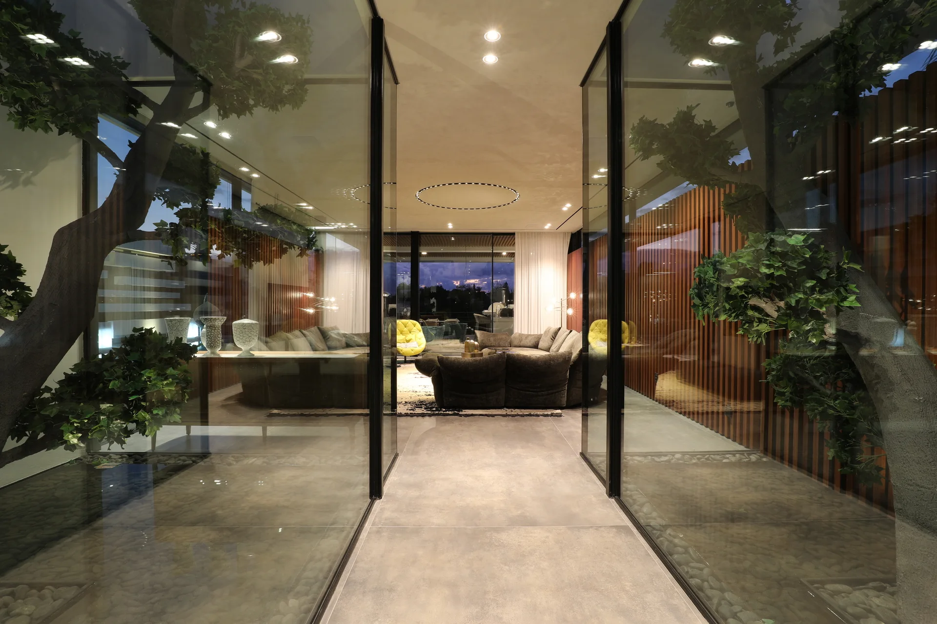 A modern corridor features large gray floor tiles and glass partitions enclosing indoor trees, with a living room visible in the background.