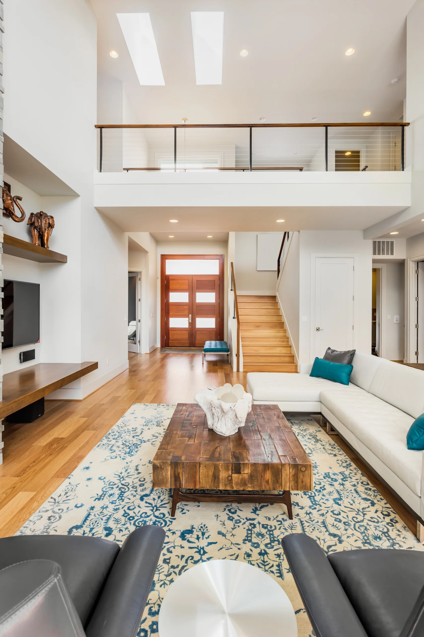A spacious living room features a white leather sectional sofa, a rustic wood coffee table, and a wooden staircase leading to a mezzanine with cable railing.