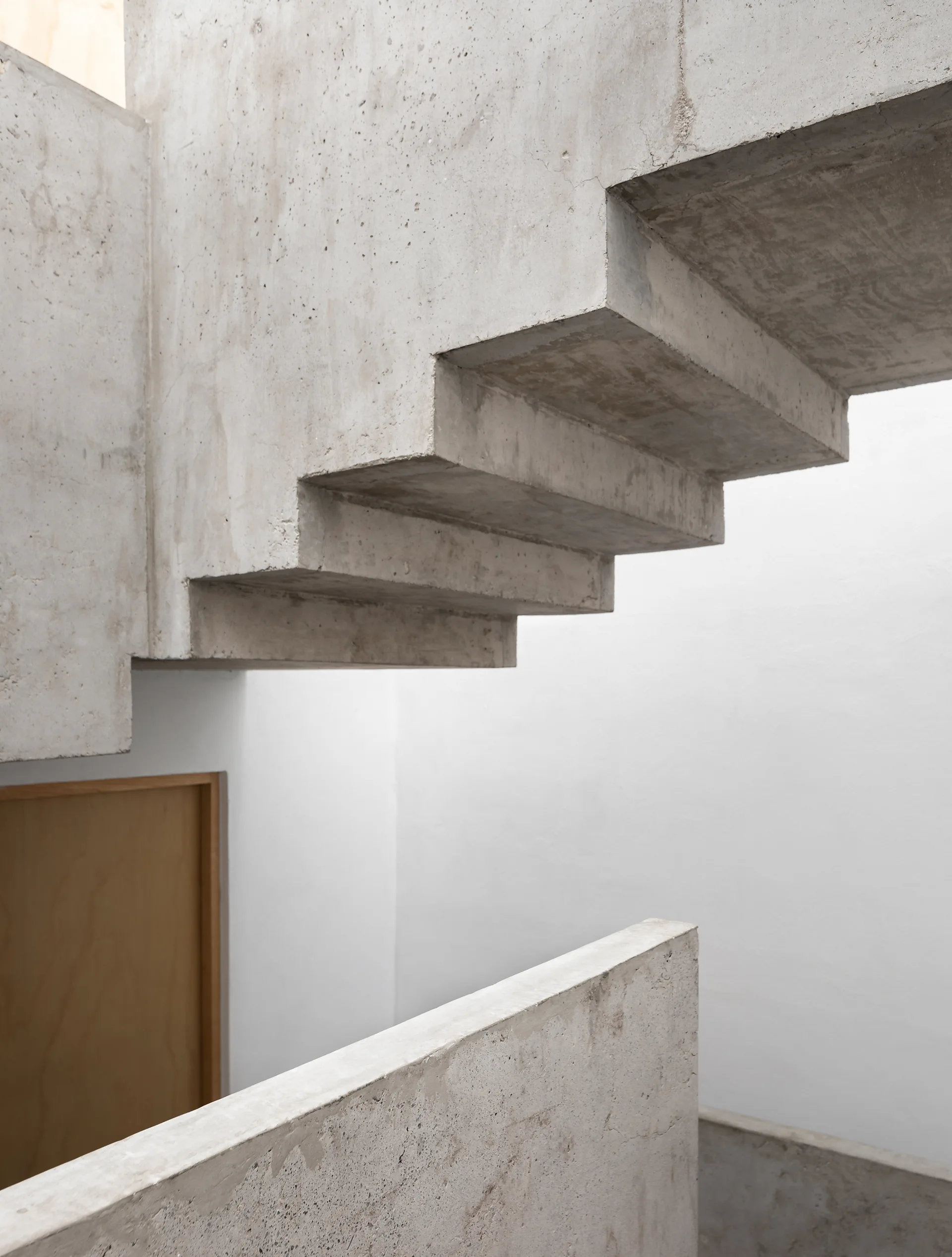 A close-up view of a minimalist concrete staircase with raw textured steps against plain white walls and a light wood door.