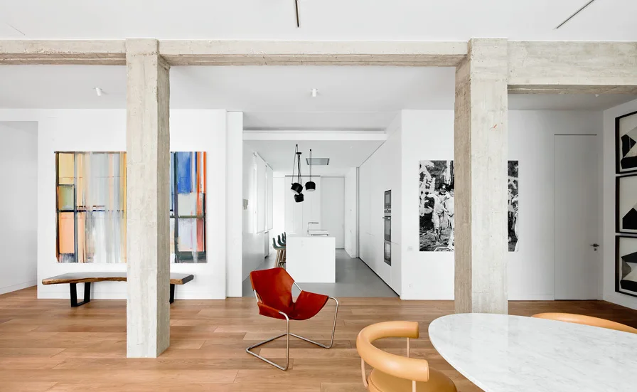 A modern open-plan living and dining space features rough concrete columns, wood flooring, a red leather chair, and minimalist white walls.