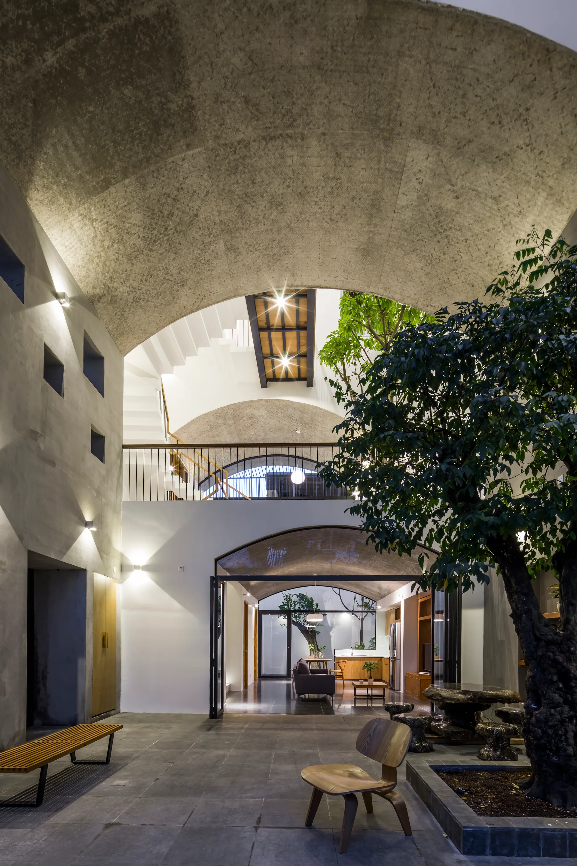 A modern indoor courtyard features a towering concrete vaulted ceiling, a mature indoor tree in a planter, a wooden chair, and a multi-level layout.