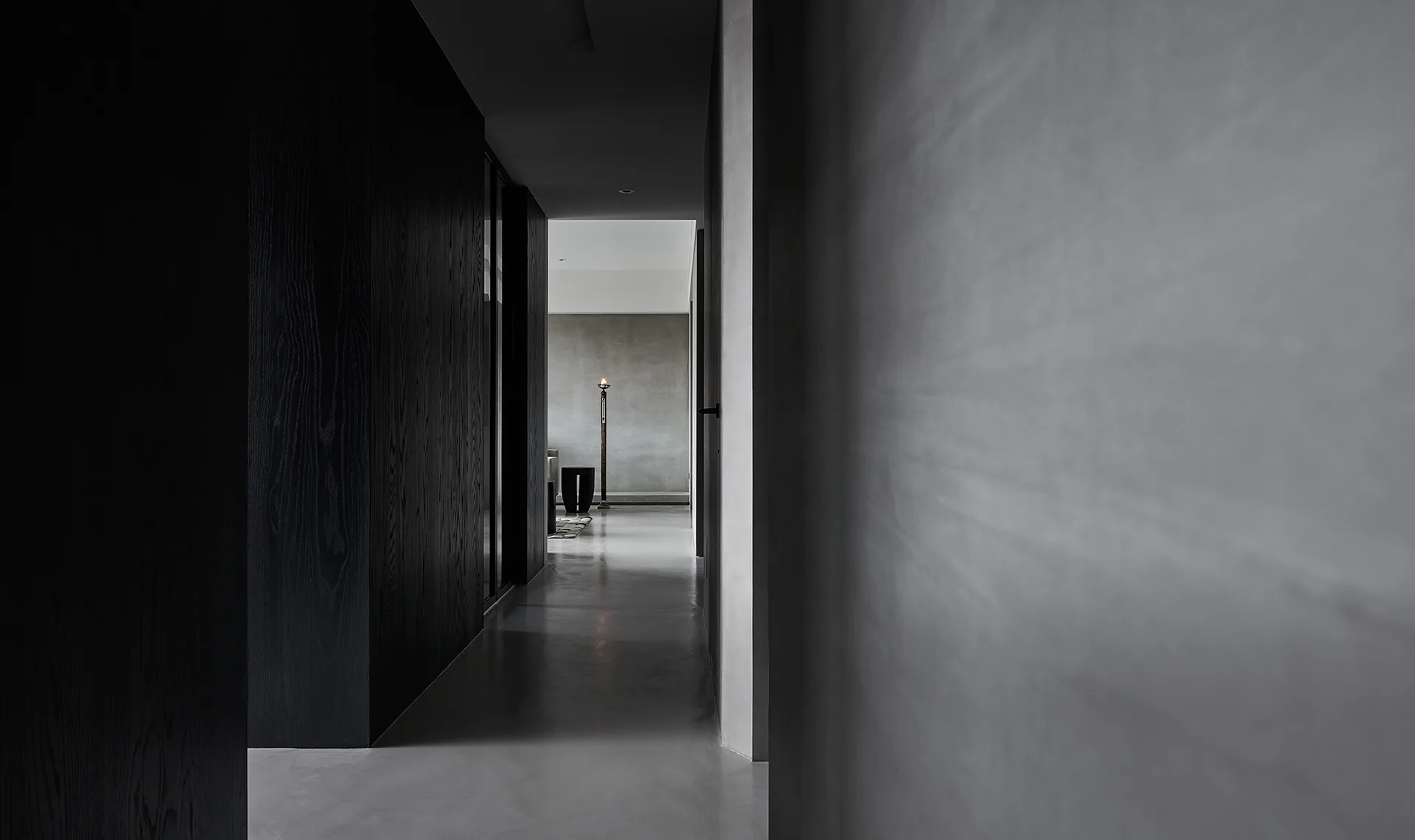 A dim minimalist corridor features charcoal wood-paneled walls and smooth concrete plaster surfaces leading to a brighter room in the distance.