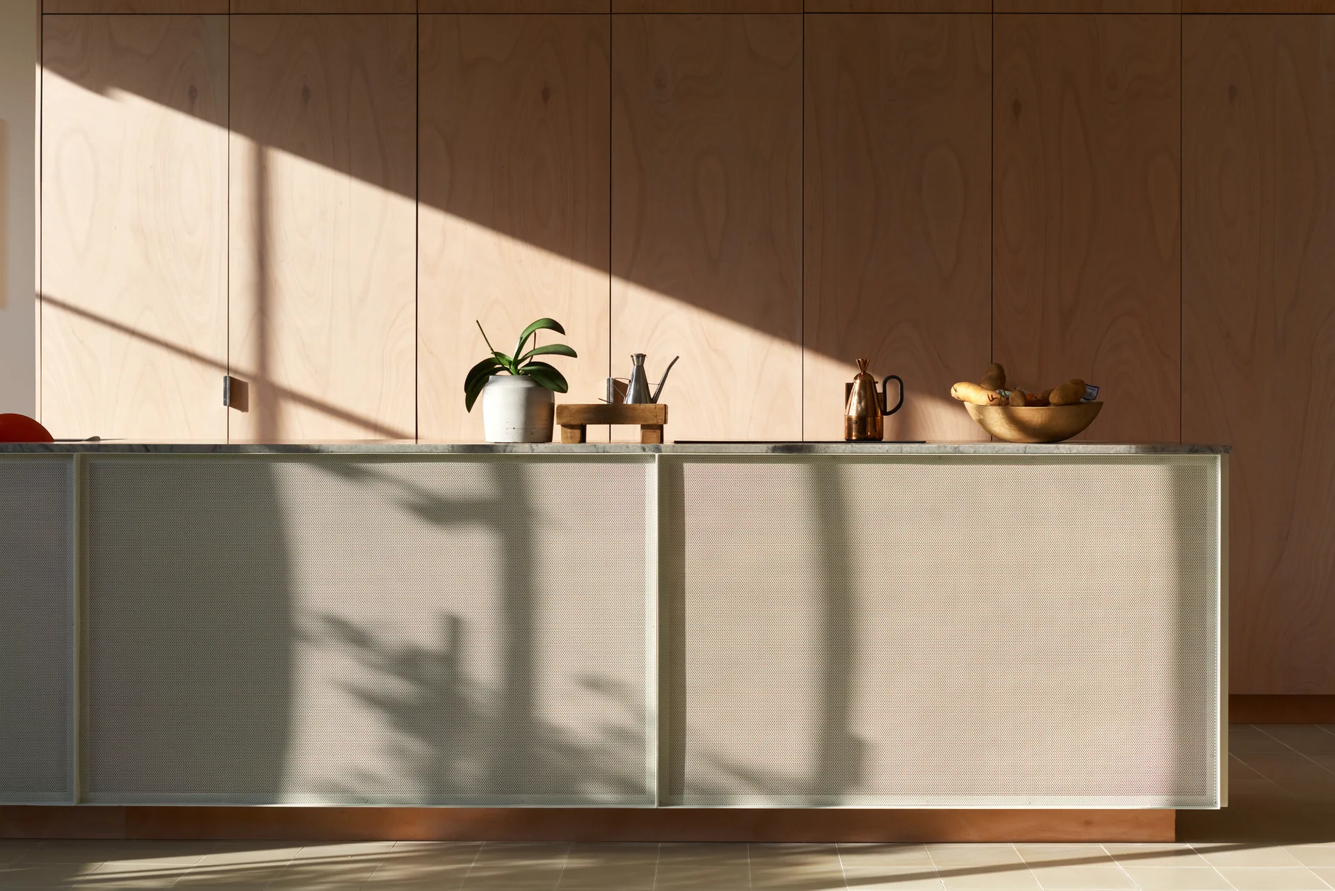 A modern kitchen island with a perforated metal front and marble countertop sits in front of a full-height wall of light wood panel cabinetry in sunlight.