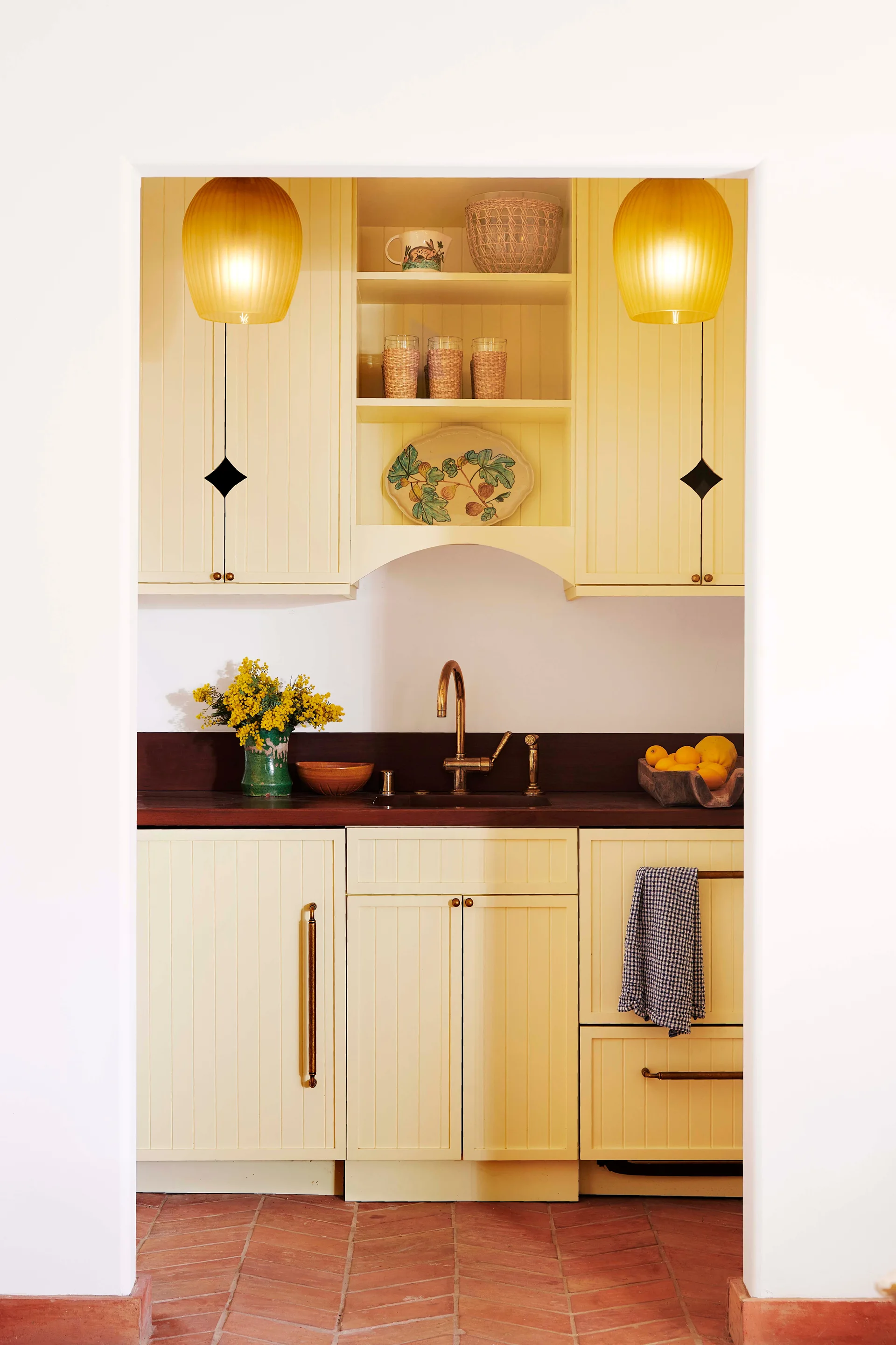 A small kitchen with pale yellow beadboard cabinetry, brass hardware, a dark wood countertop, and red terracotta floor tiles.