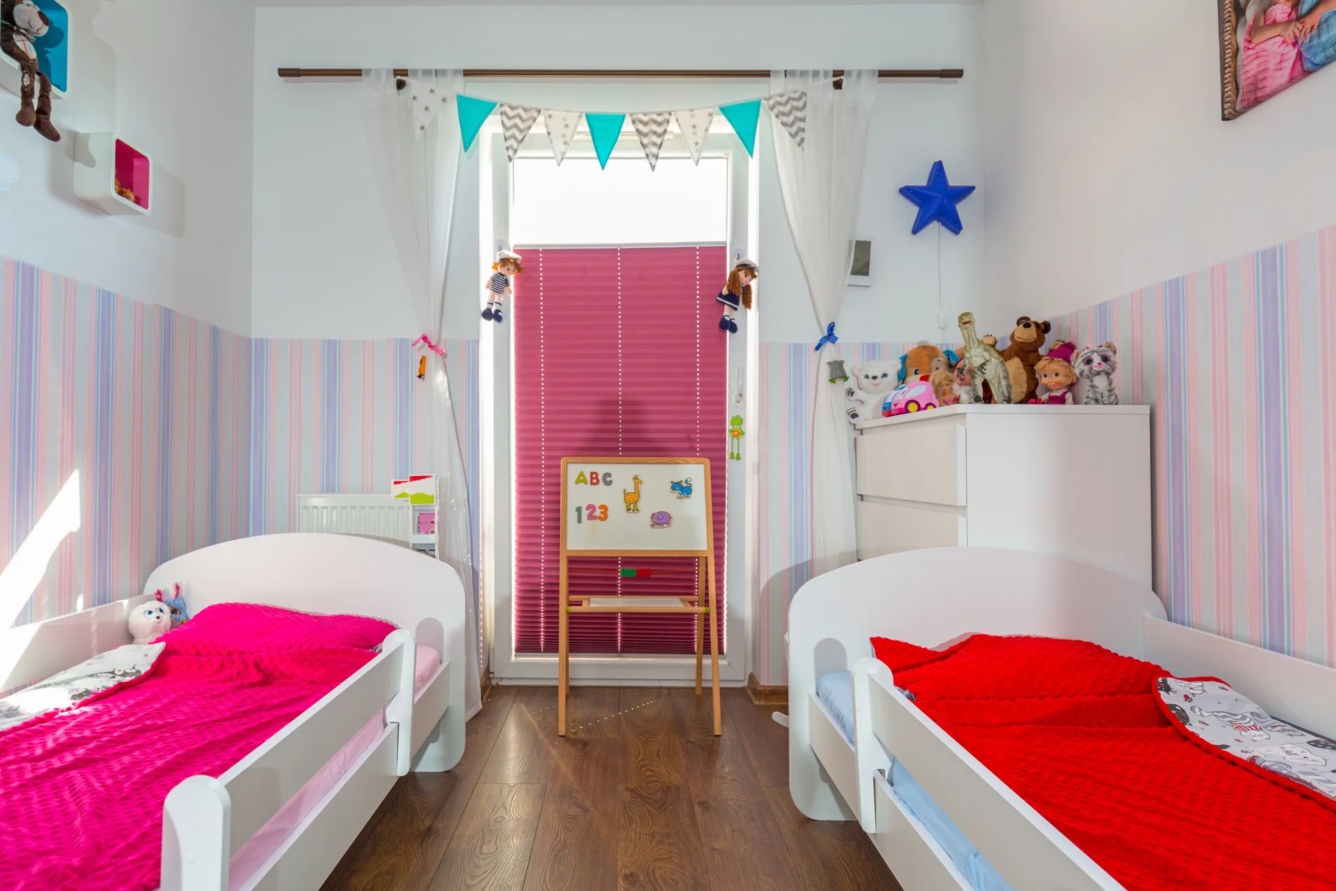 A shared children's bedroom features two white twin beds, pastel striped wallpaper, and a central window with bright pink blinds.