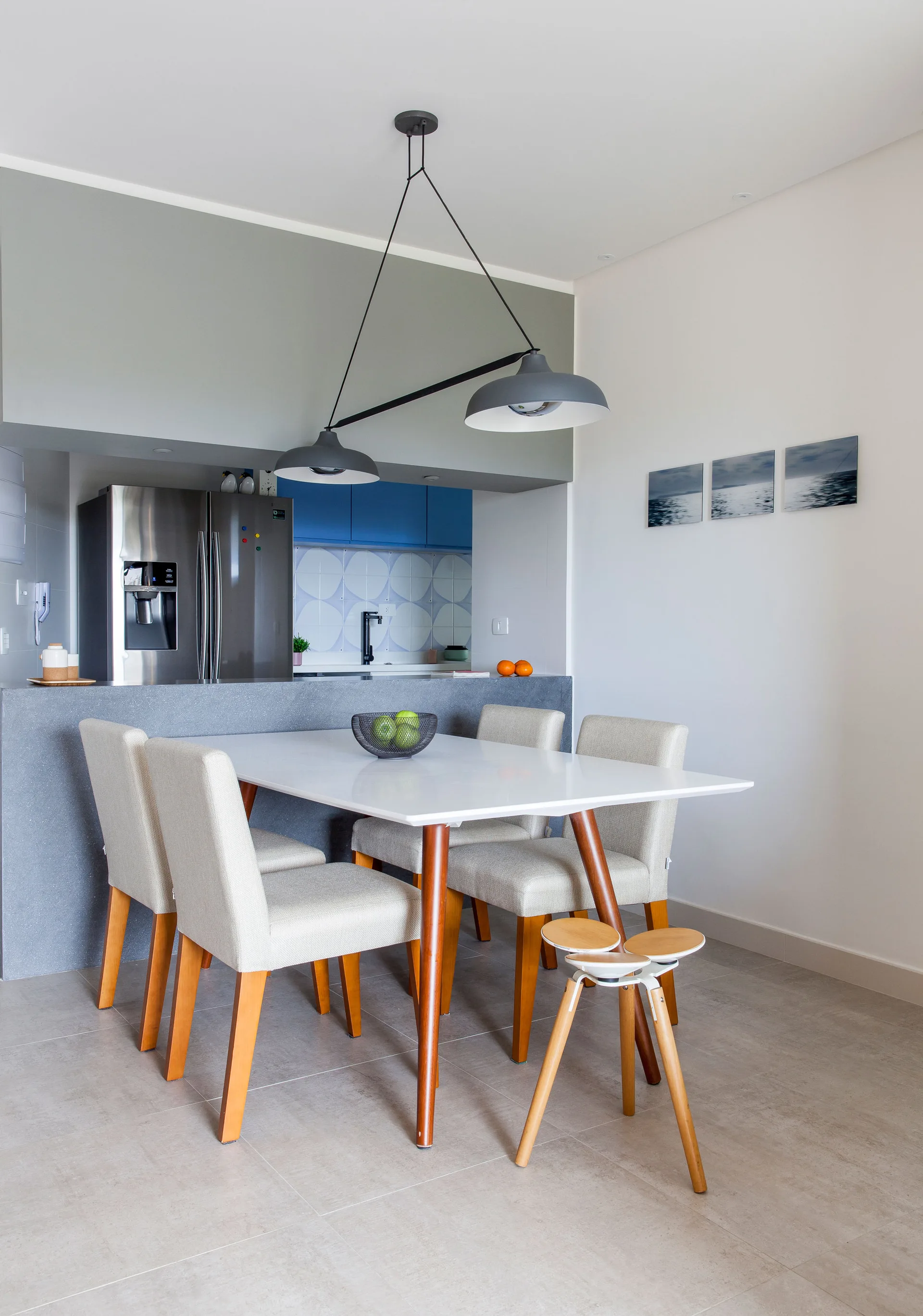 A modern dining area features a white wood-legged table, upholstered chairs, a low double pendant light, and a stone kitchen peninsula.