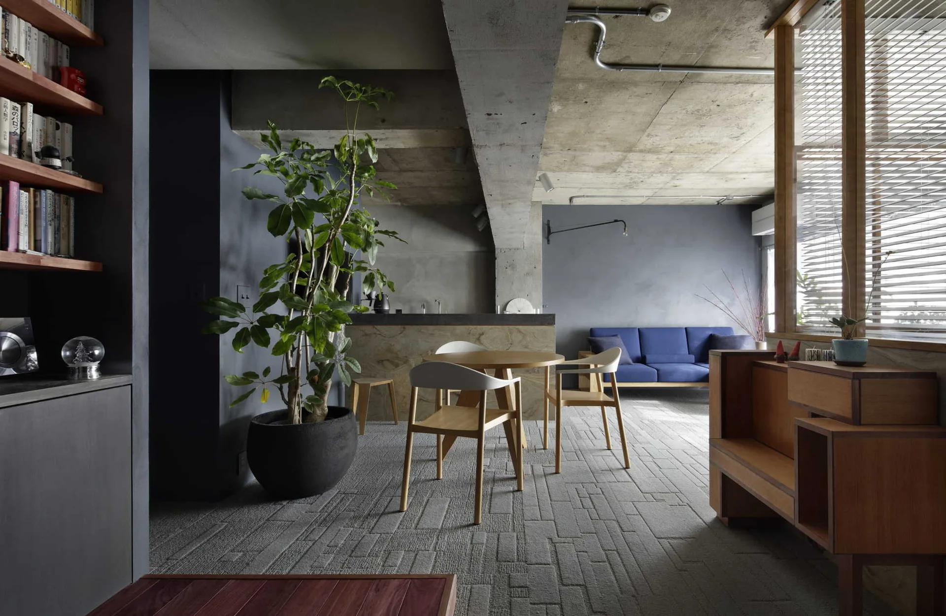 A modern living space features an exposed concrete ceiling, a round wood dining set, a large potted plant, and a blue sofa.
