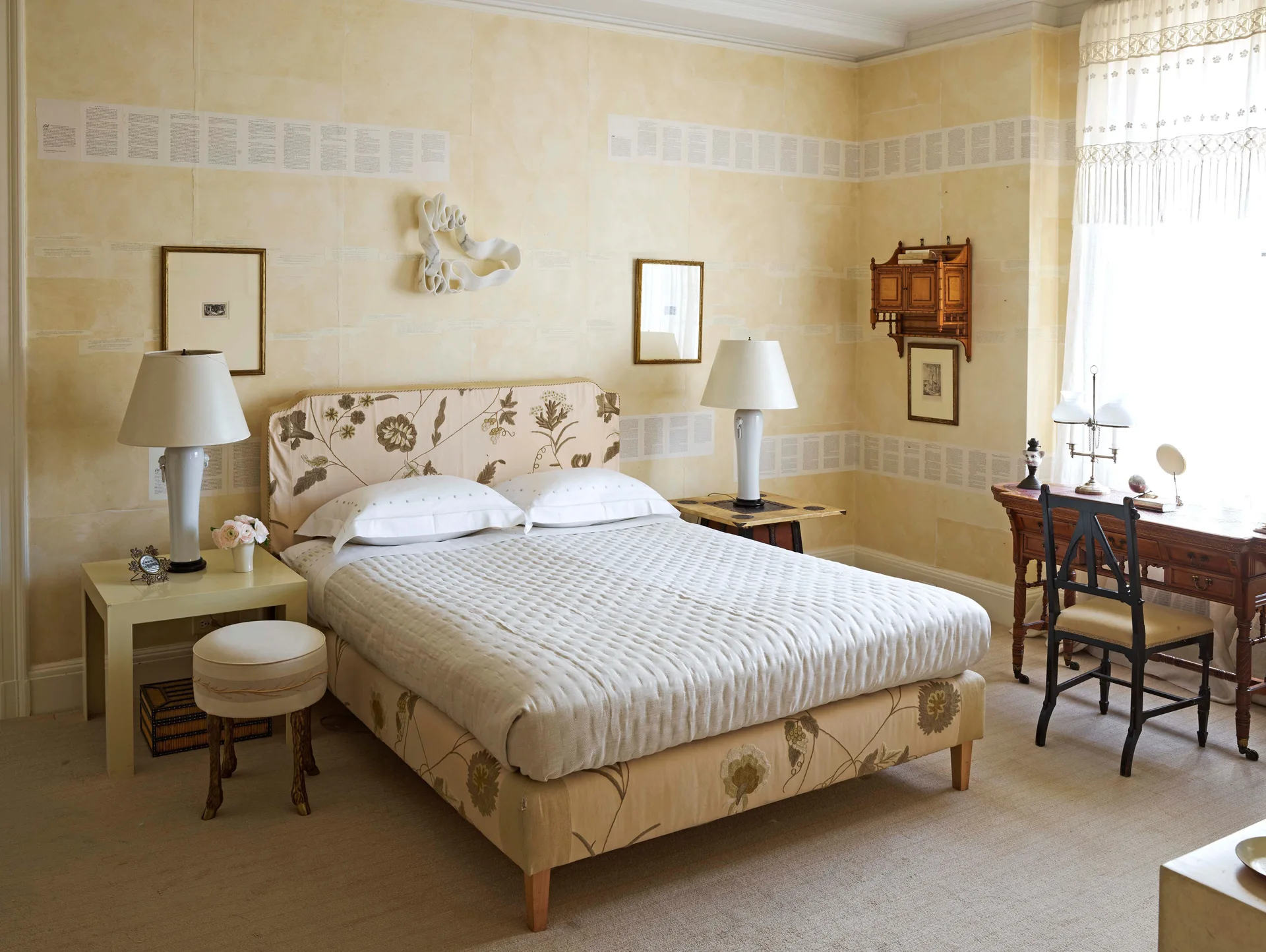 A yellow bedroom features a floral upholstered bed, matching white table lamps on nightstands, a wooden writing desk, and patterned wall borders.
