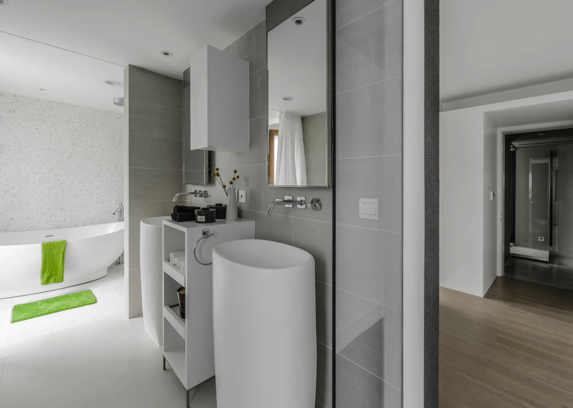 A modern grey tiled bathroom featuring two white cylindrical pedestal sinks, rectangular mirrors, and a white freestanding bathtub in the background.