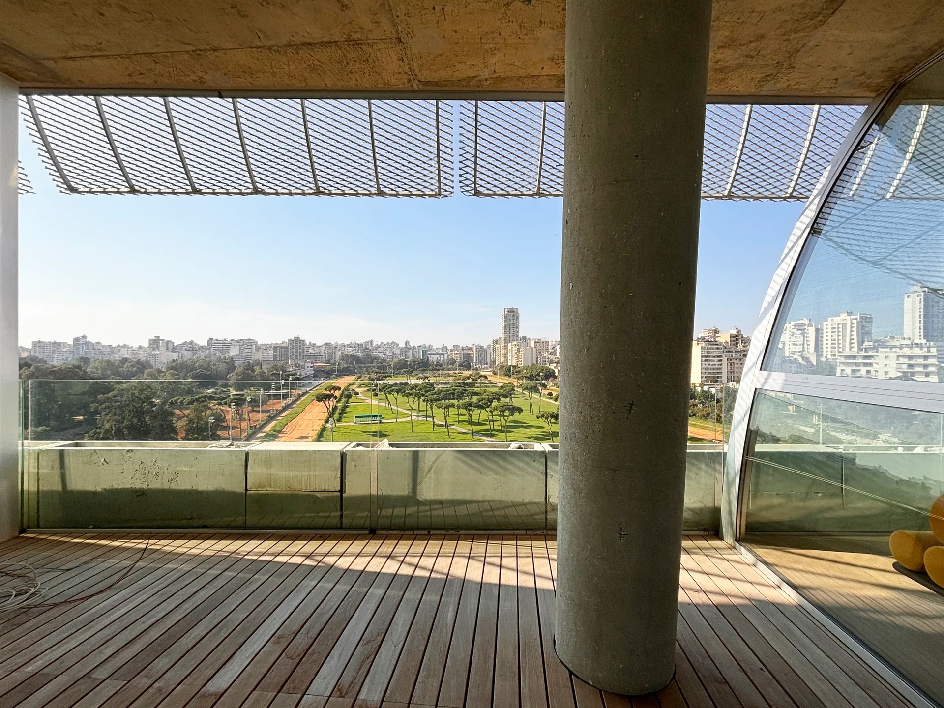 A modern outdoor terrace with wooden decking, a large round concrete column, and a clear glass railing overlooking an expansive city park.