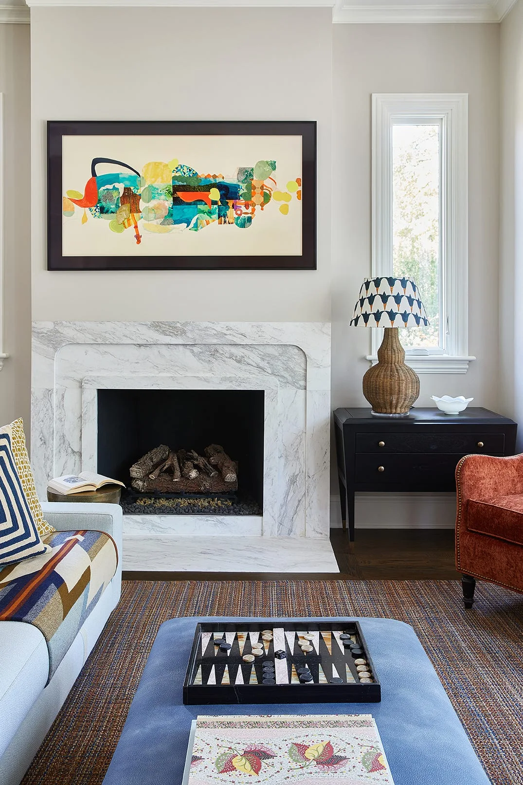 A living room features a veined white marble fireplace, a black framed abstract art piece, a wicker lamp, and a rust-colored velvet armchair.
