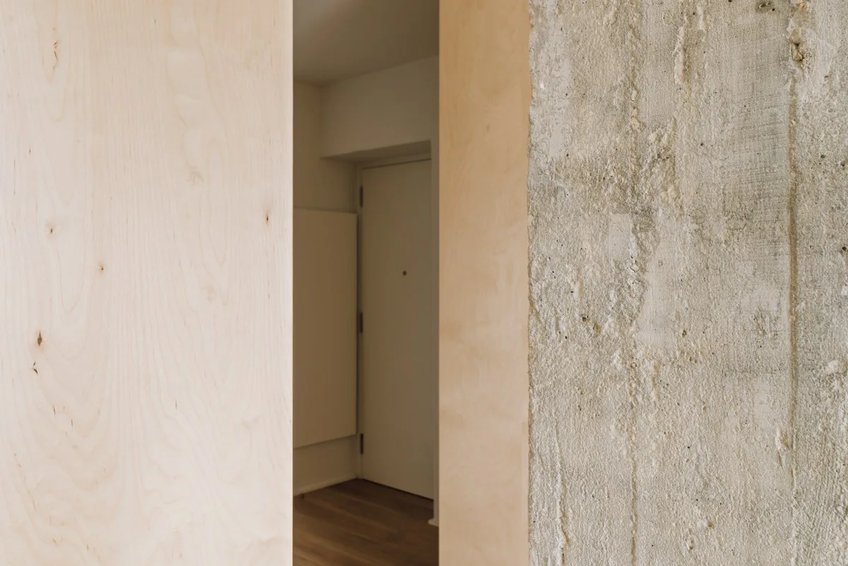 A close-up view of a doorway transition framed by light wood plywood paneling on one side and a textured raw concrete wall on the other.