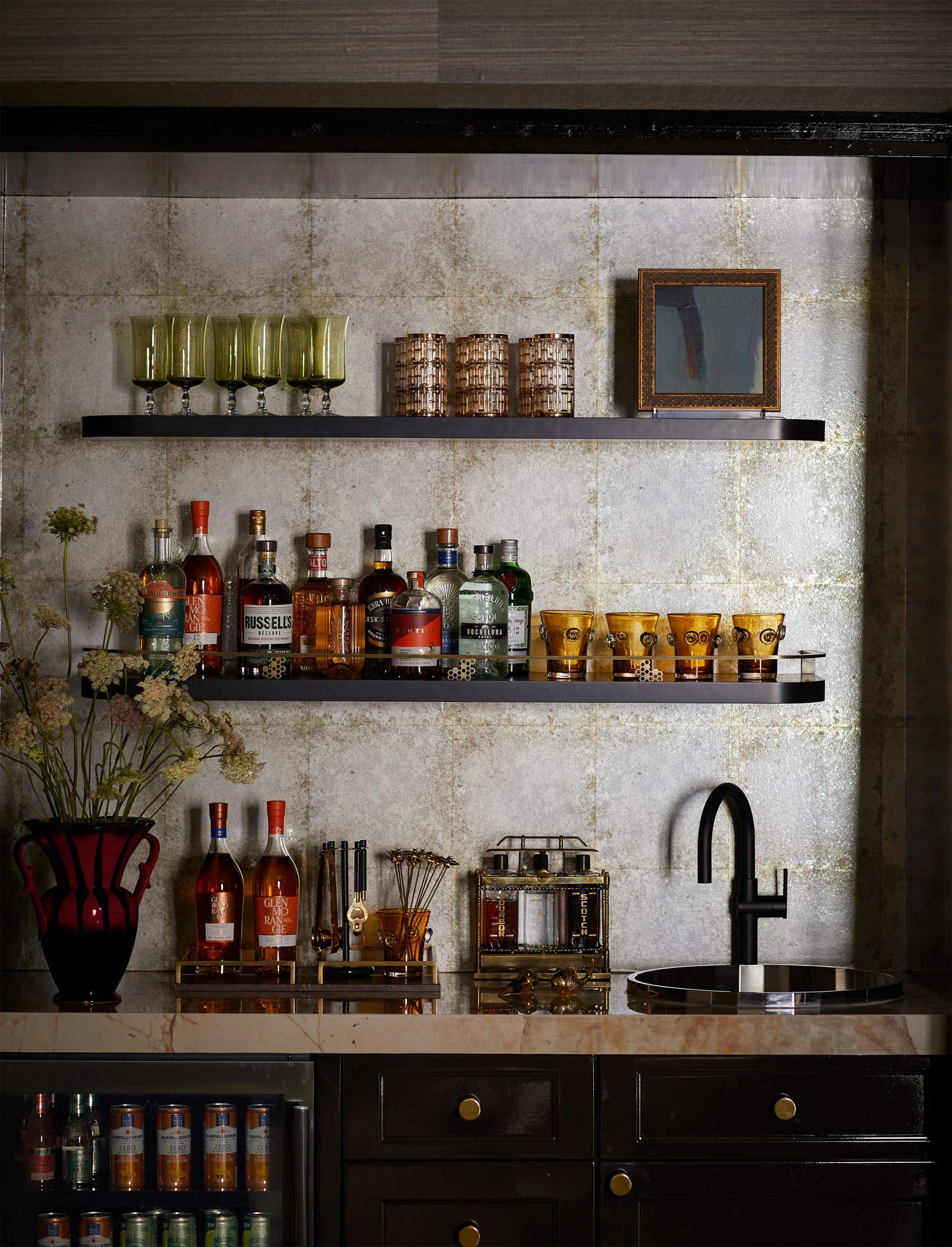 A home bar features black floating shelves, amber glassware, a black faucet, and a reflective gold-toned tiled backsplash.
