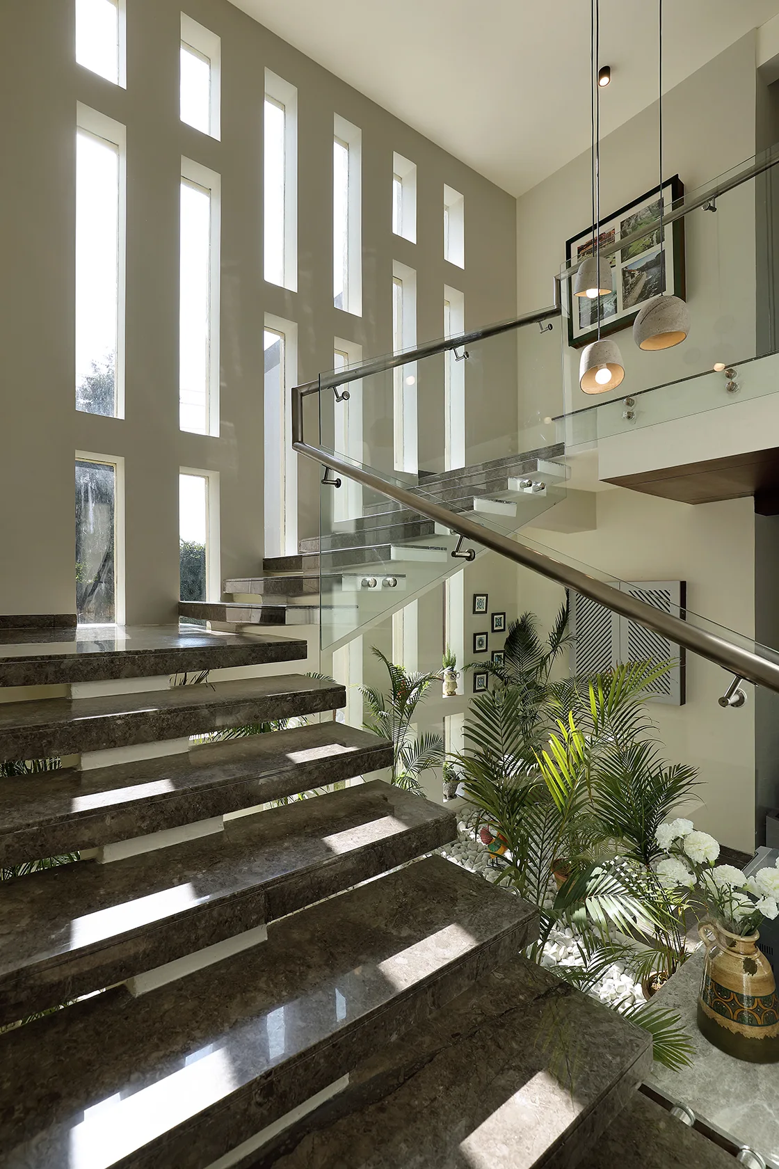 A contemporary staircase features polished marble treads, glass railing, a rhythmic pattern of tall narrow windows, and an indoor garden underneath.
