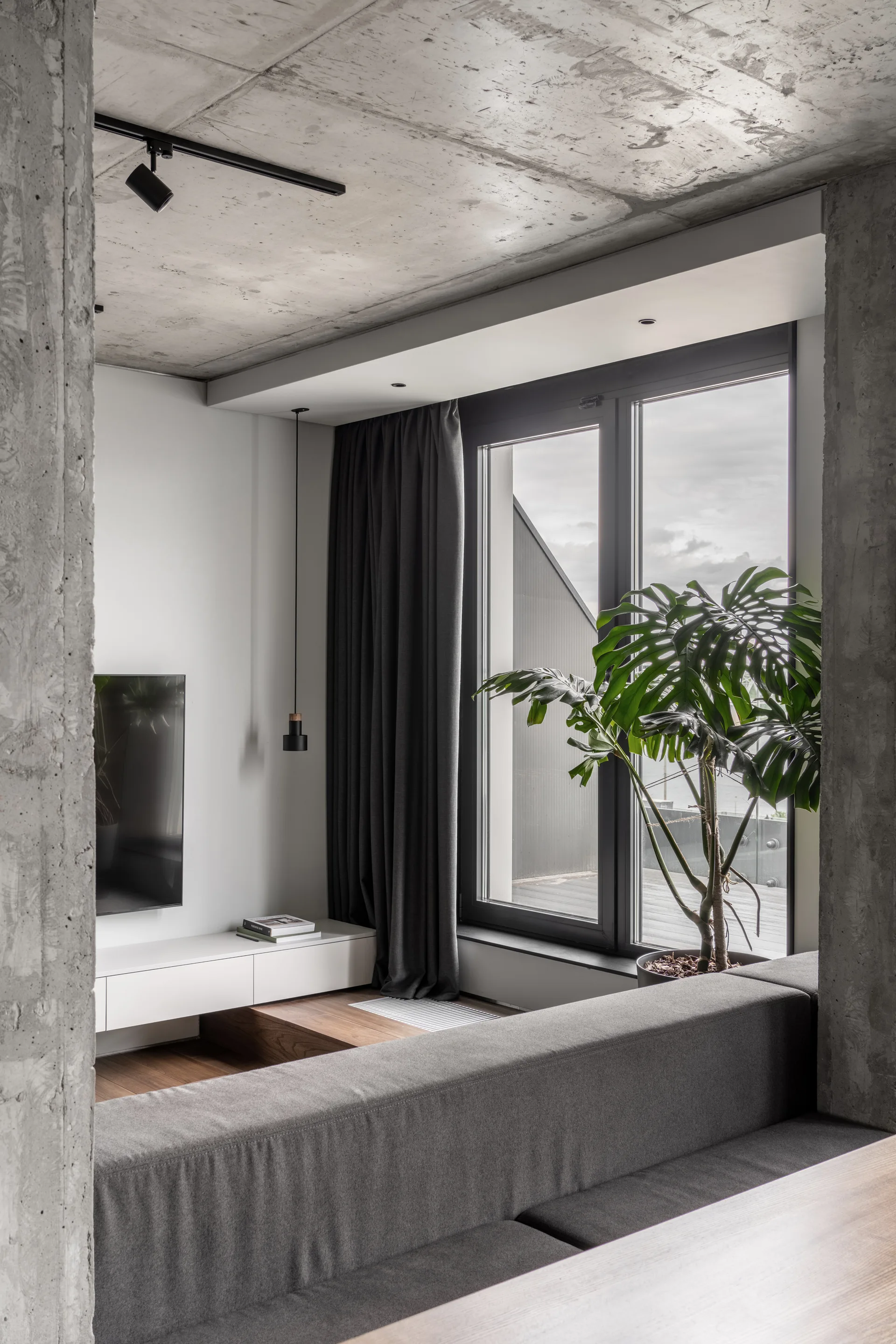 A minimalist living room features an exposed concrete ceiling, a low white media console, a grey sectional sofa, and a large window.