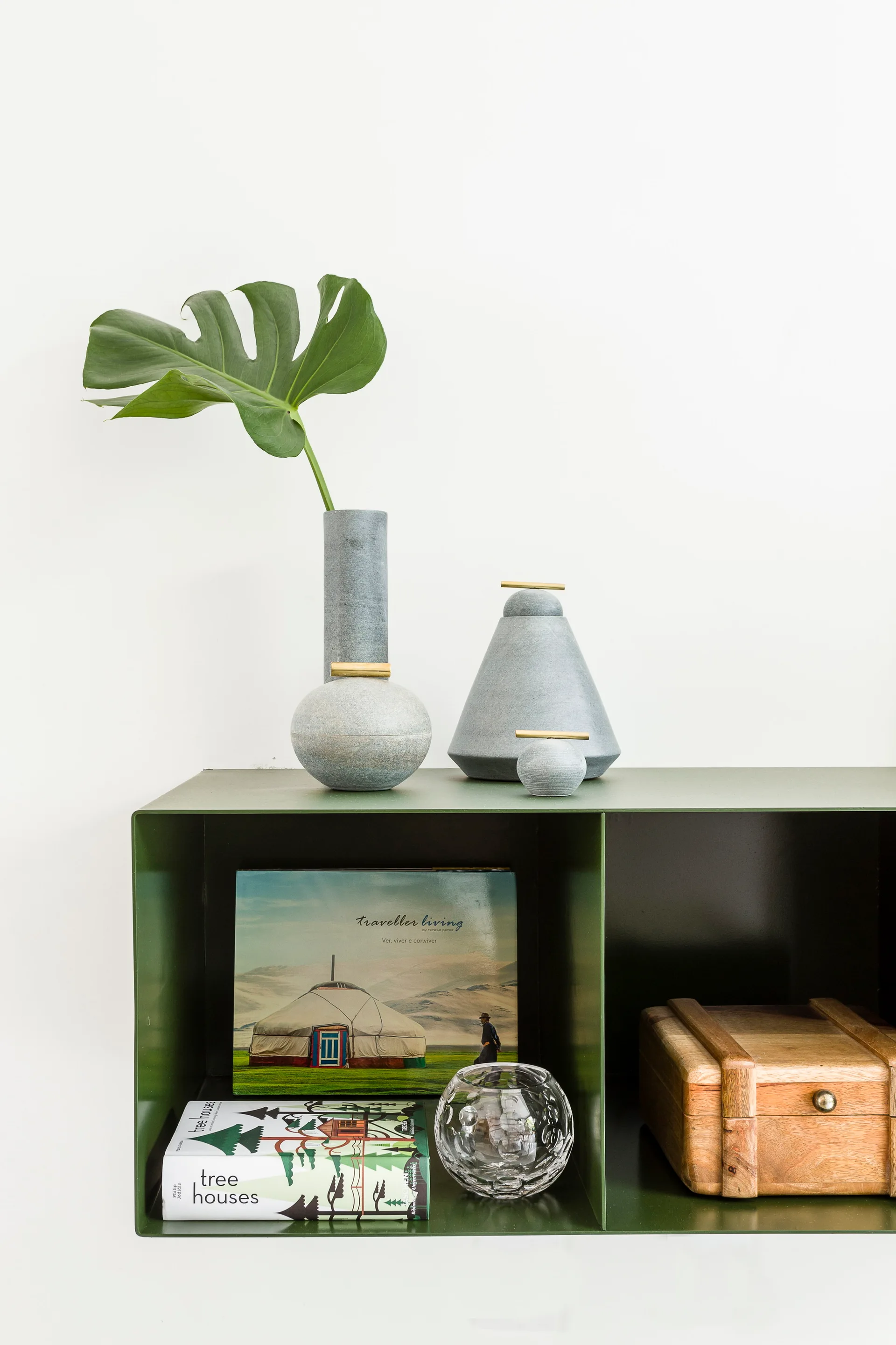 A green metal wall shelf holds books, a wooden box, and a glass bowl, topped with three grey stone vases and a single green monstera leaf.