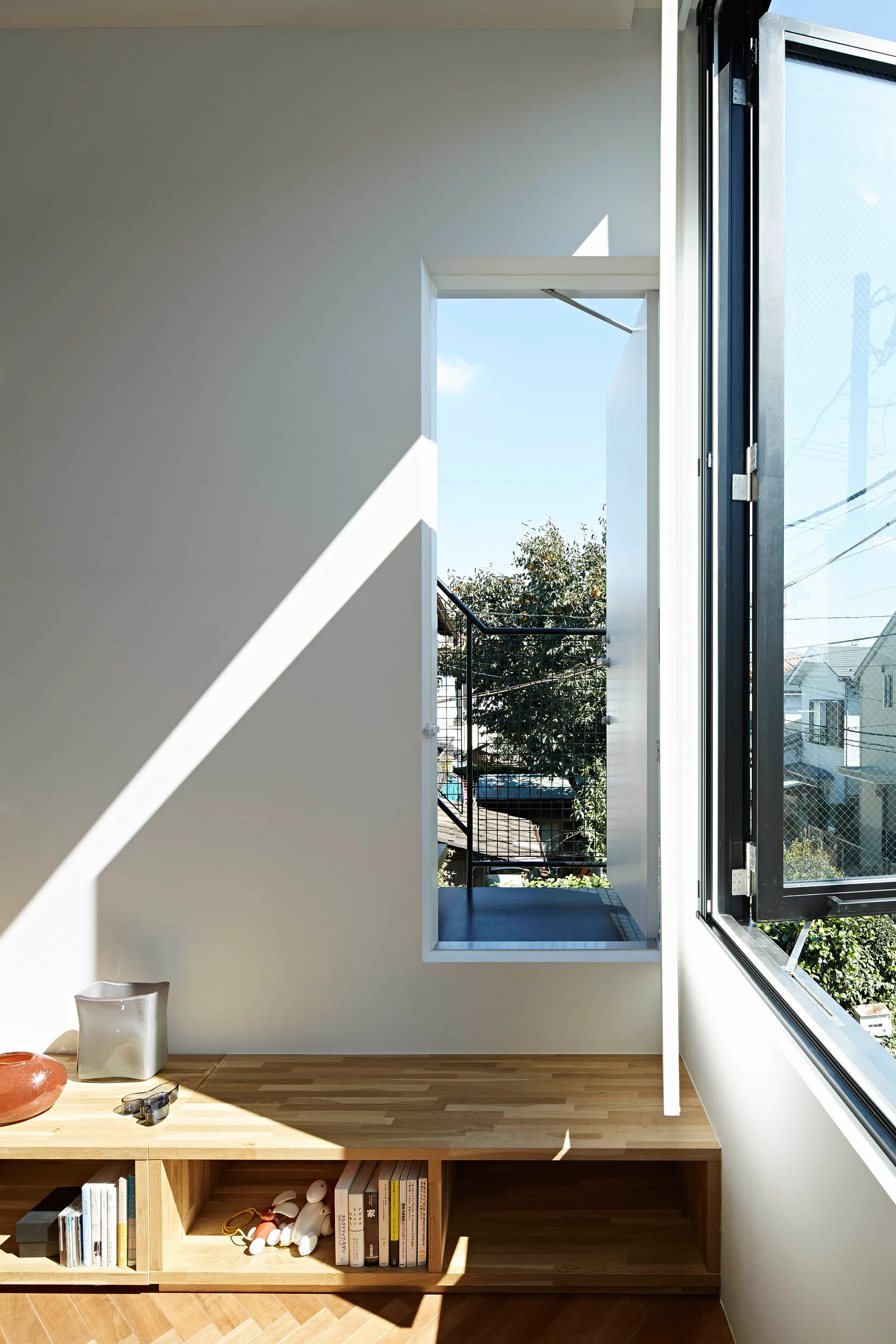 A low light-wood bookshelf sits under a white wall featuring a narrow open doorway to a balcony and a large window casting diagonal sunlight.