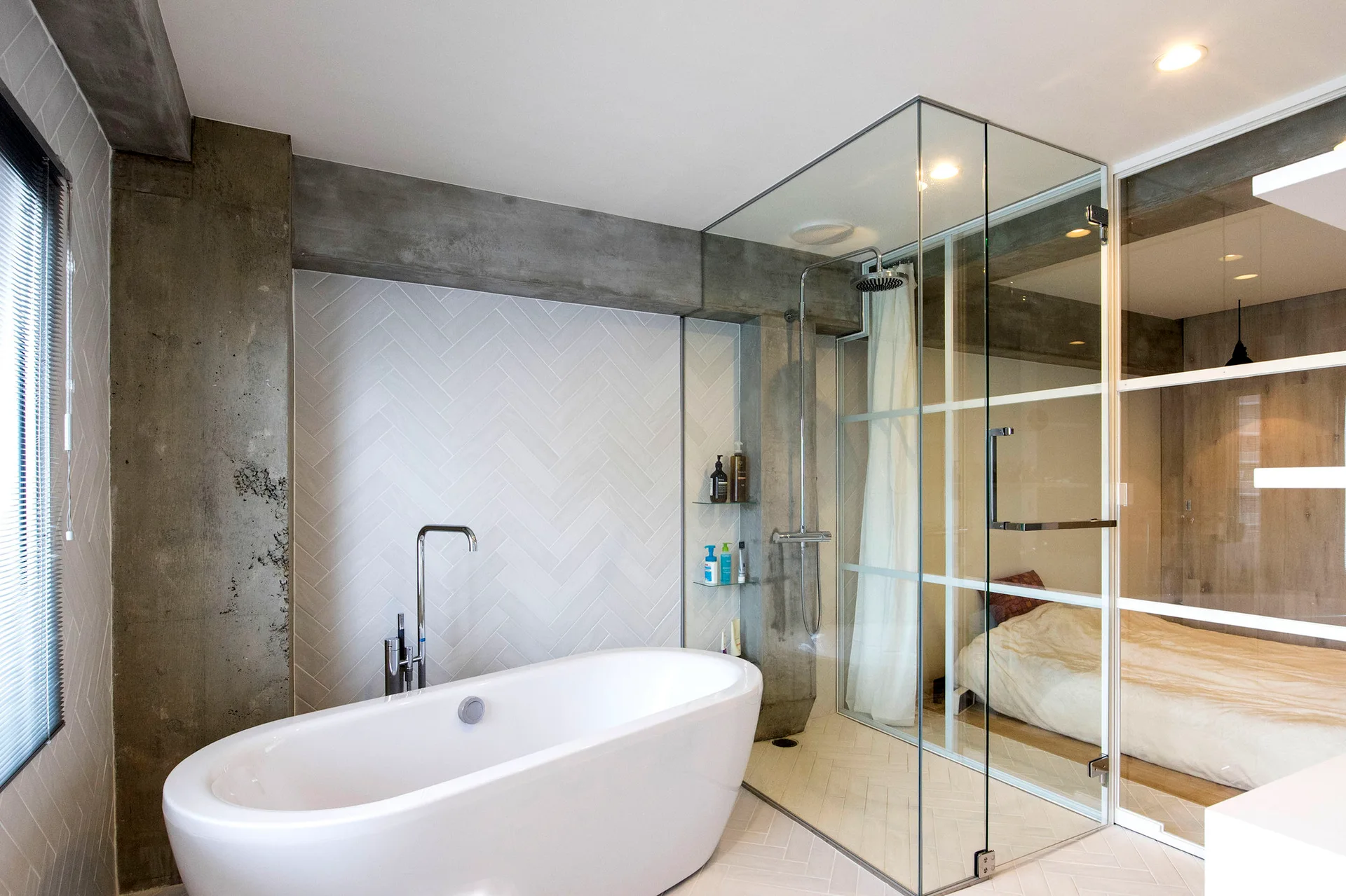 A modern bathroom featuring a white freestanding bathtub and a glass-enclosed shower next to an exposed concrete beam and a glass bedroom wall.