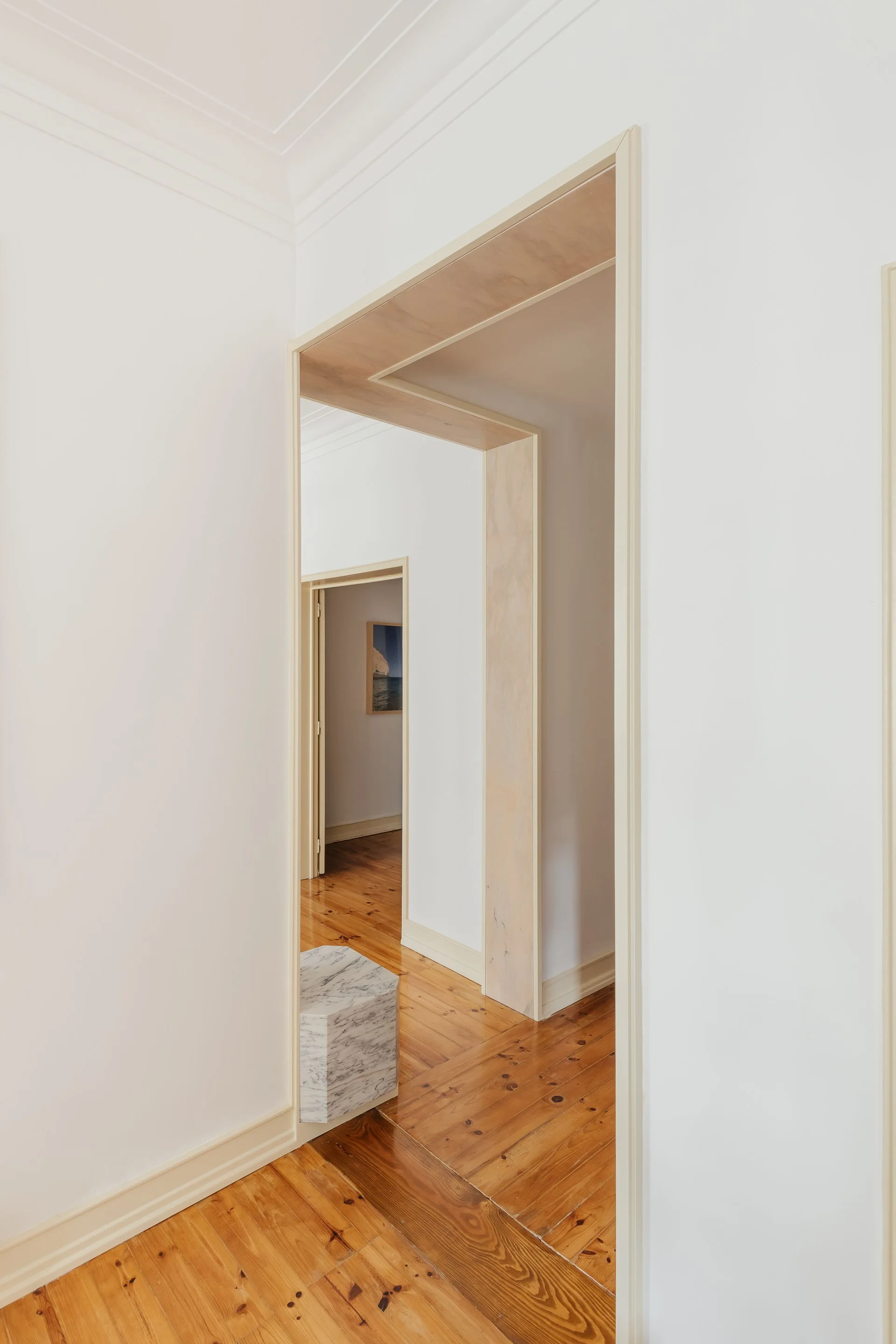 A bright white hallway with natural hardwood floors features two doorways framed in light-colored wood casing and a small hexagonal marble plinth.