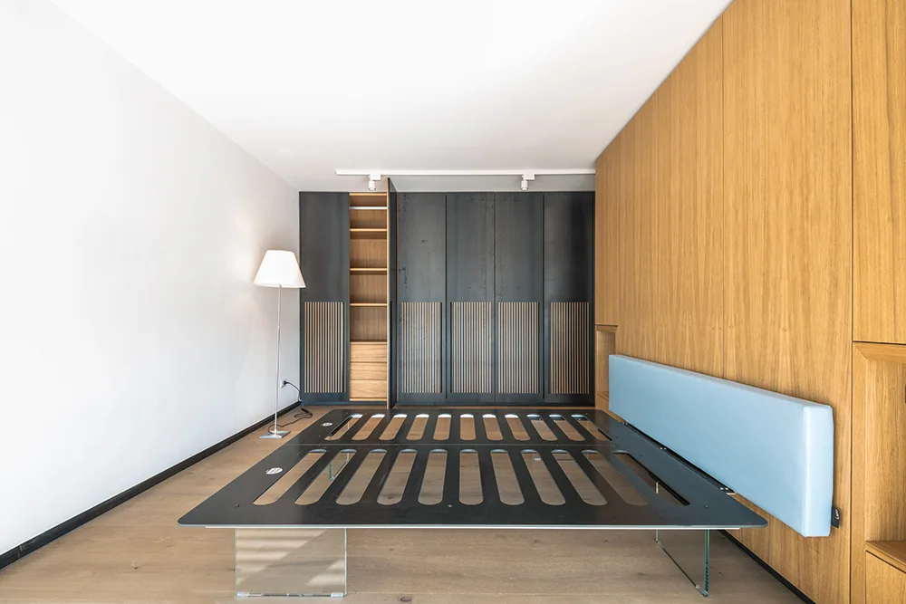 A minimalist bedroom featuring a black bed frame on transparent glass legs against a wood paneled wall and dark slatted wardrobe.