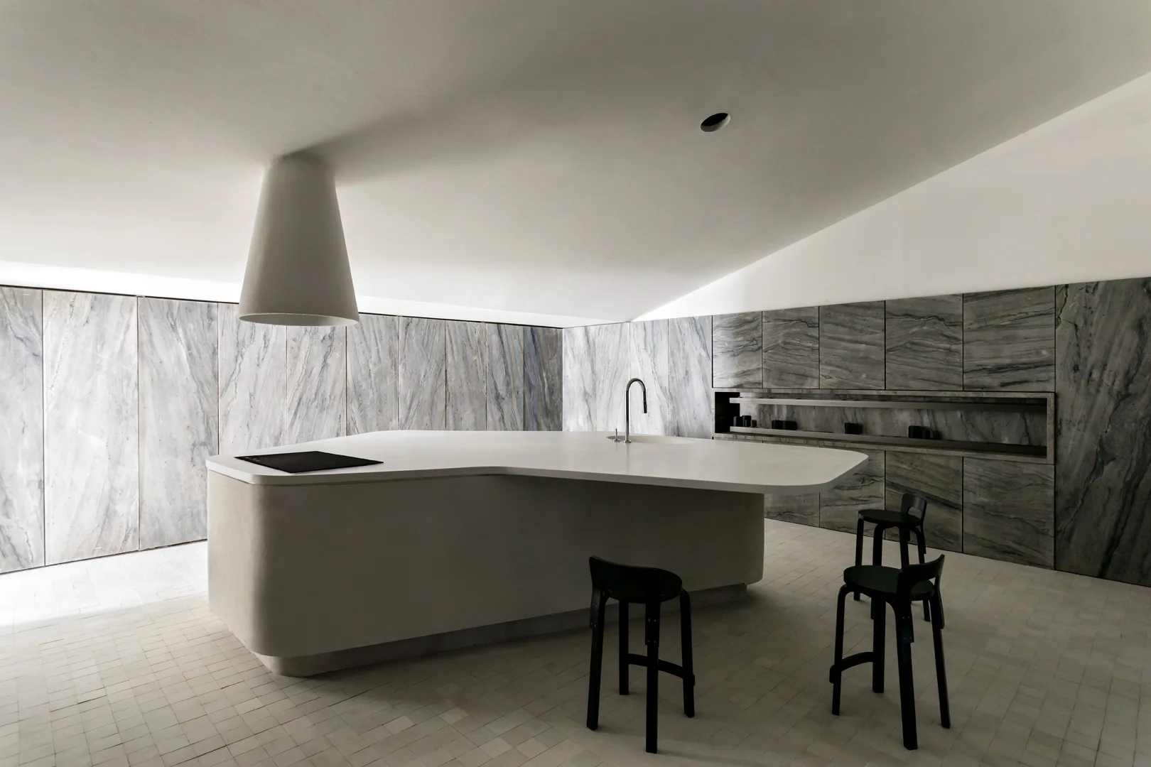 A minimalist kitchen features a large curved white island with black stools, a conical range hood, and grey veined stone paneling on the walls.