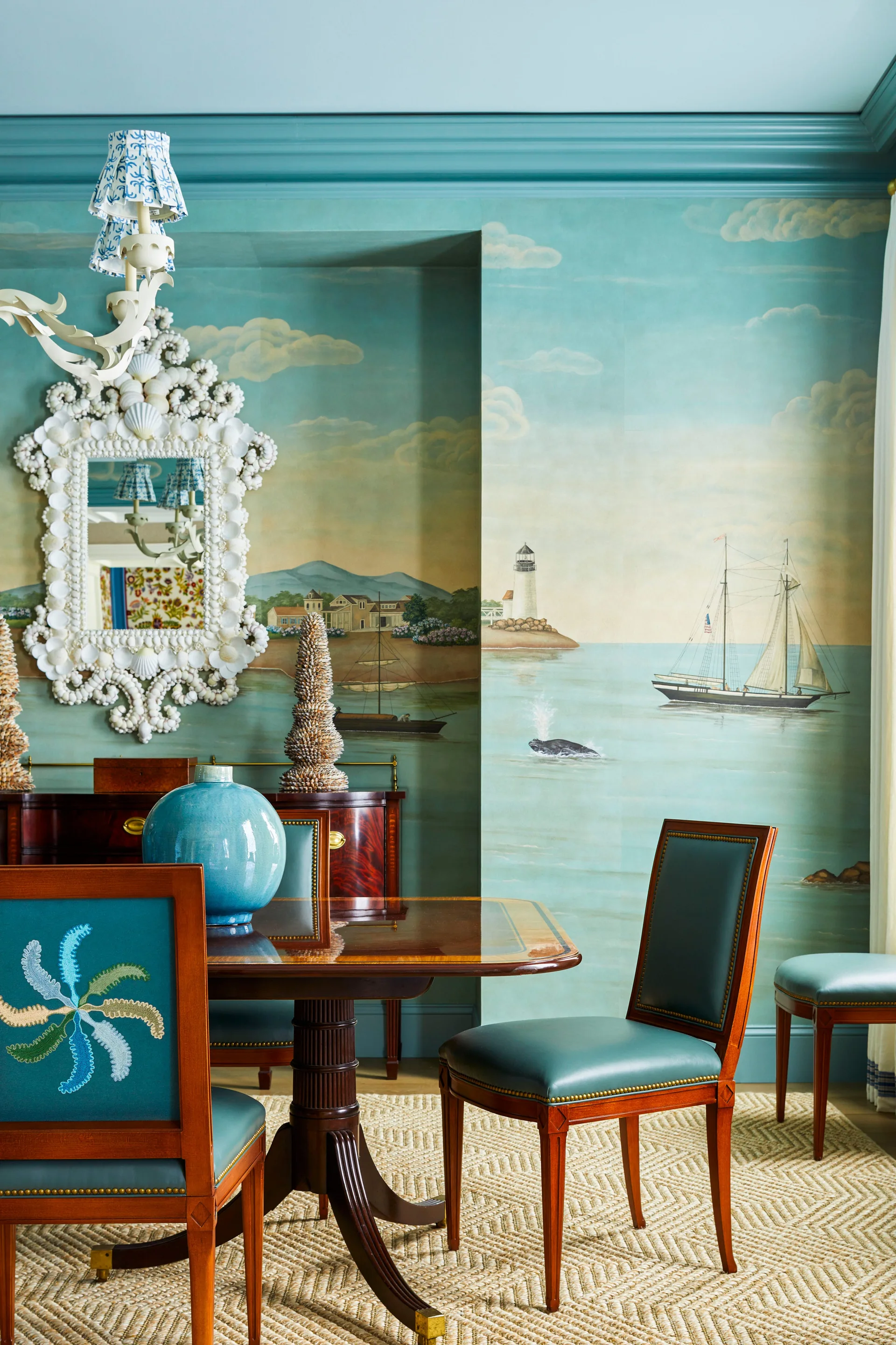 A dining room features a coastal landscape wall mural, a white shell mirror, a dark wood dining table, and teal upholstered chairs on a woven rug.